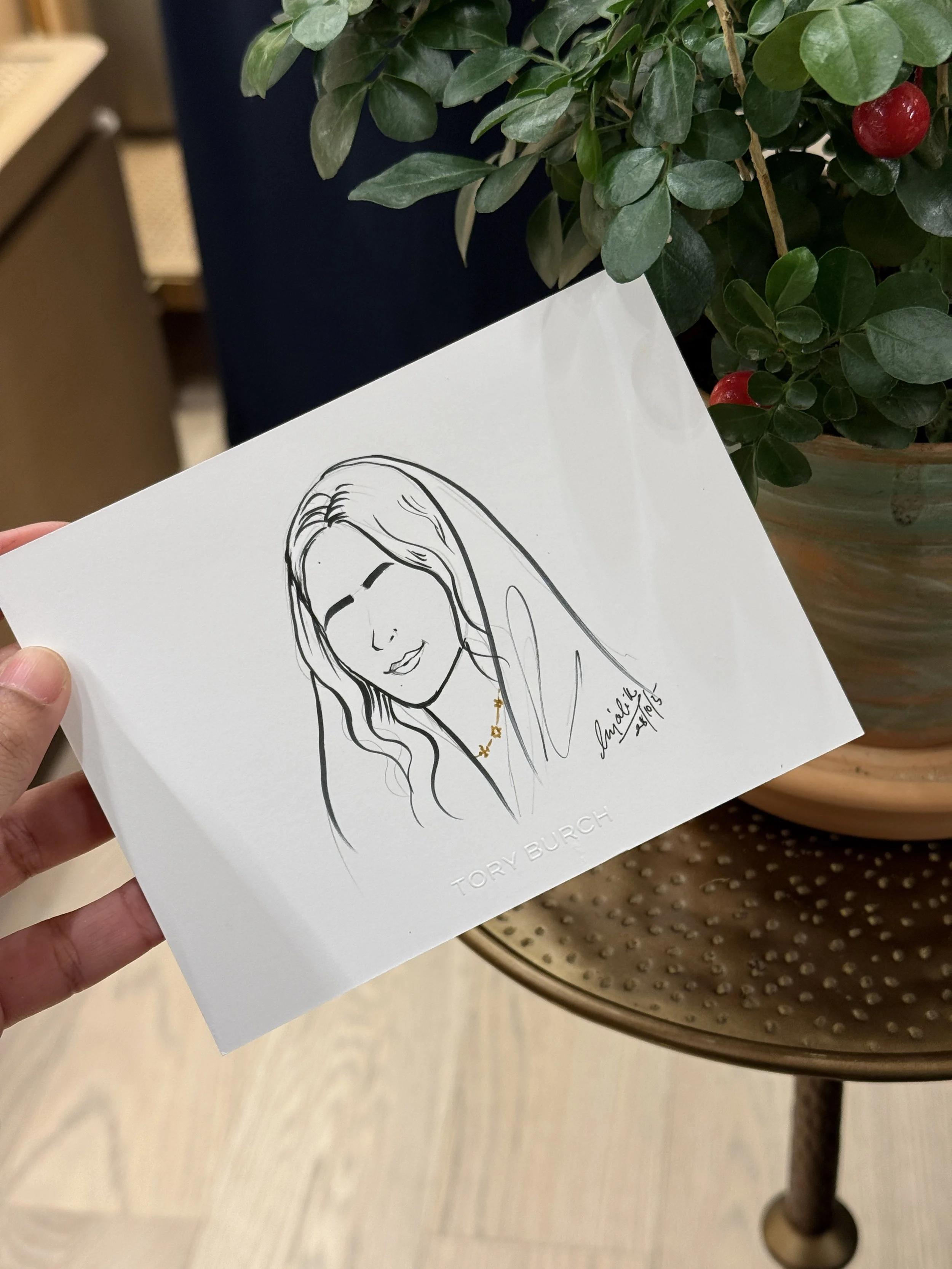A hand holding a Tory Burch promotional card featuring a line drawing of a woman's face, with the Tory Burch logo and signature, in front of a potted plant with red berries.