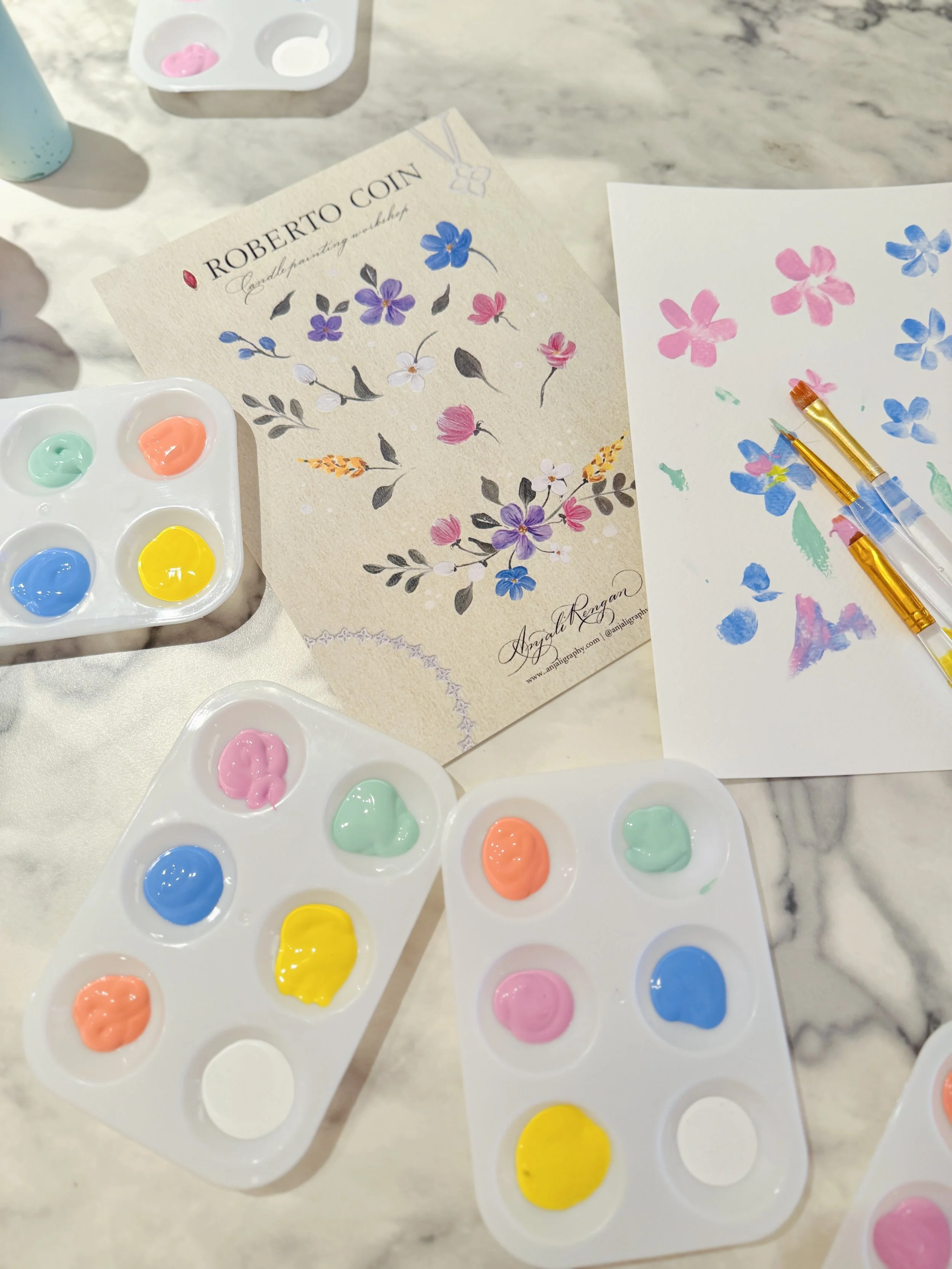 Watercolor paint palette with pink, blue, yellow, green, and orange paints, watercolor paper with painted flowers, and small watercolor paint pots on a marble surface.
