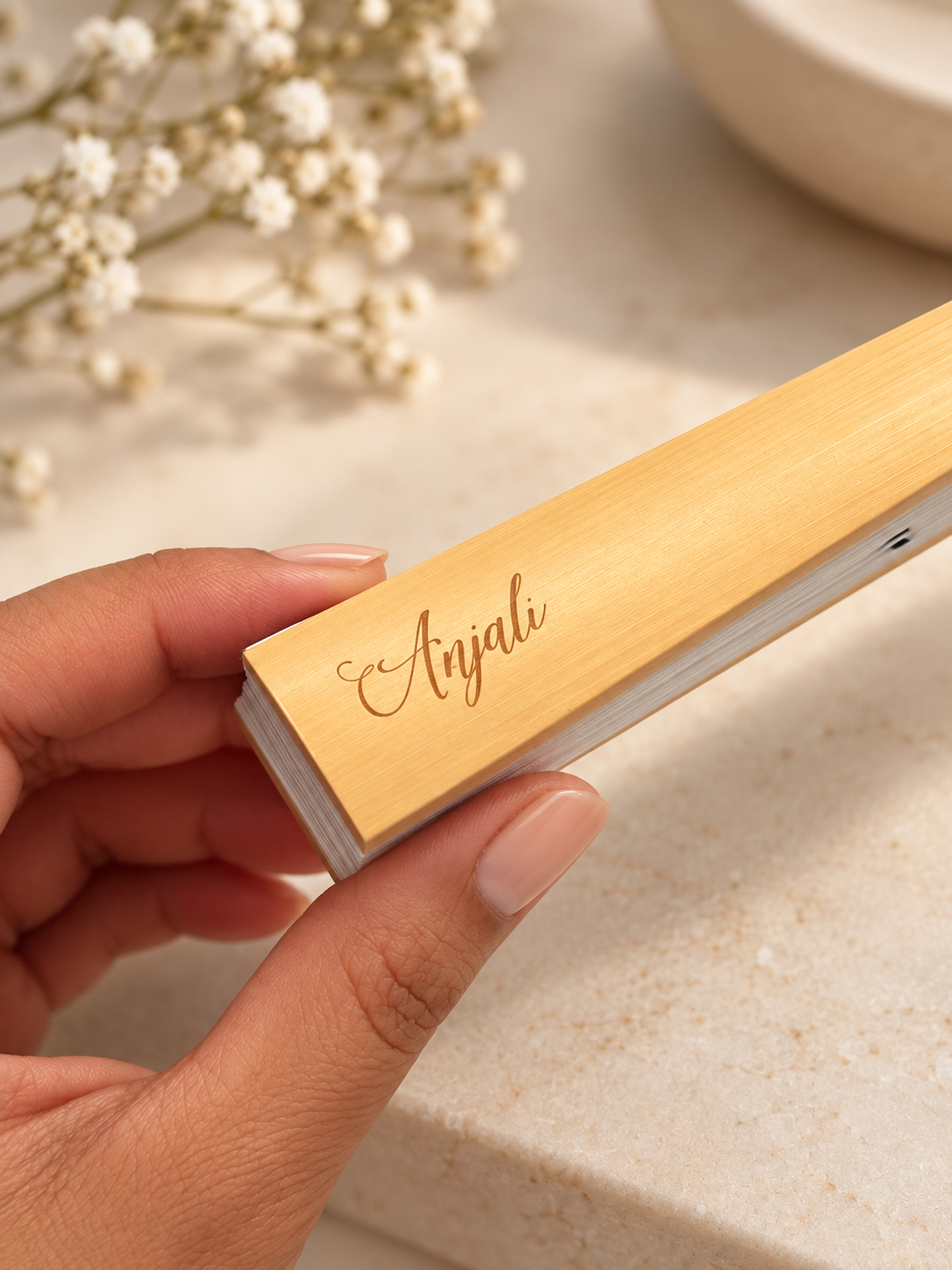 A hand holding a small card with the name 'Anjali' written in cursive, against a background of white flowers and a beige surface.