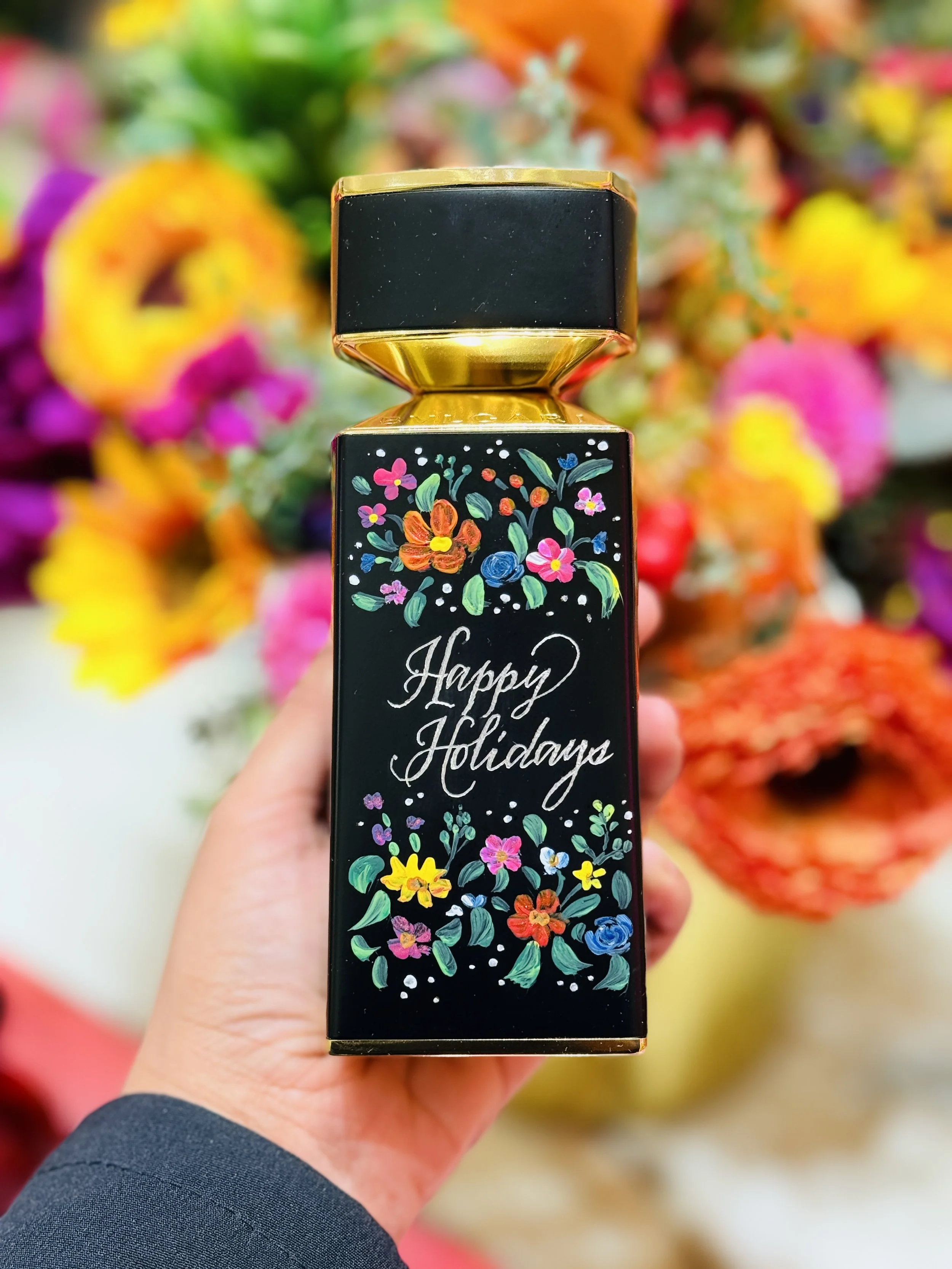A hand holding a black rectangular box with colorful floral illustrations and the words "Happy Holidays" on it, with a blurred background of vibrant flowers.
