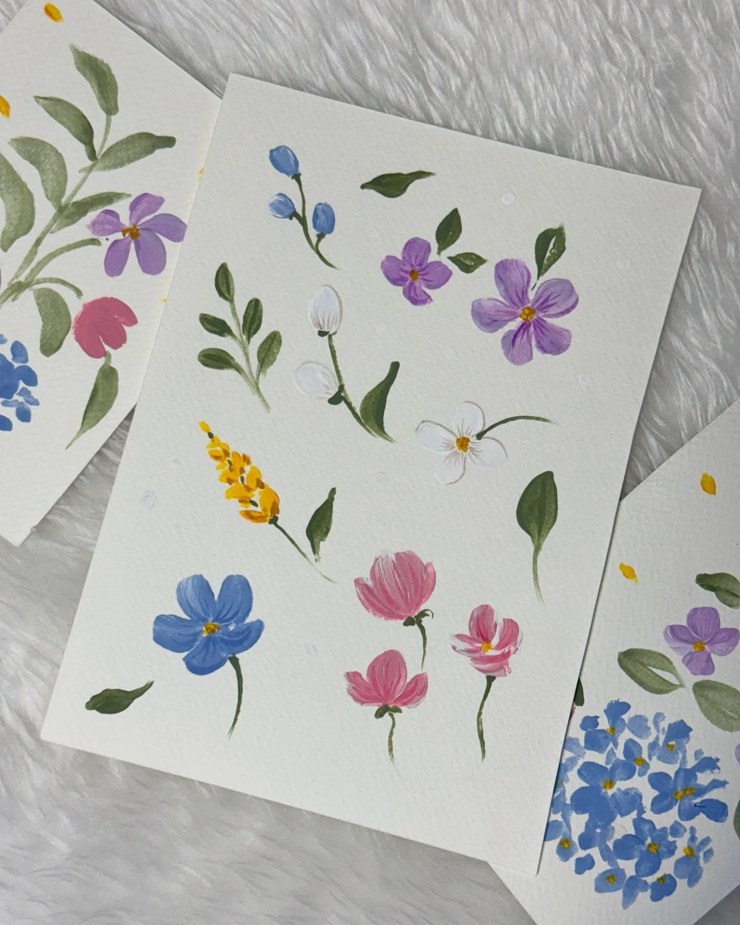 Several watercolor floral illustrations featuring various flowers in shades of purple, pink, blue, yellow, and white, with green leaves, on textured watercolor paper.
