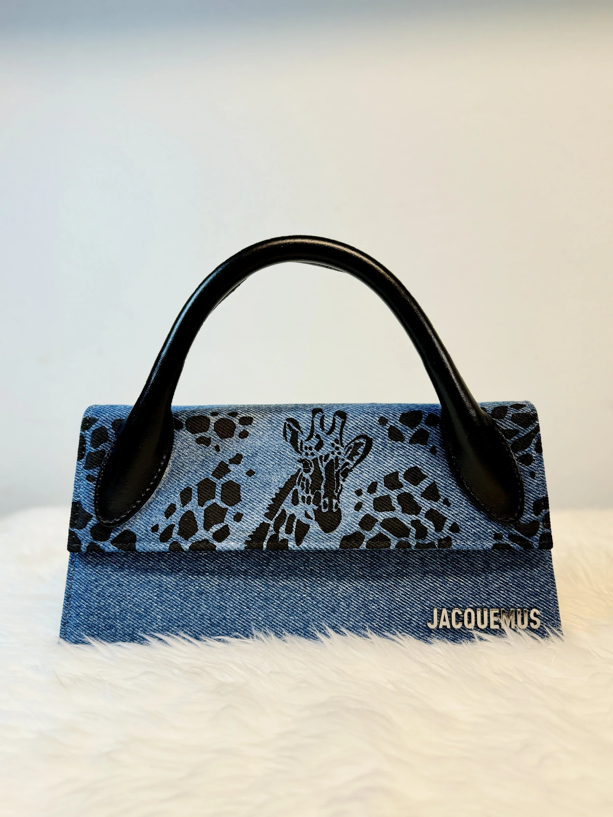 Jacquemus designer handbag with a denim body and black handles, featuring a black giraffe silhouette and the brand name 'JACQUEMUS' on the bottom right corner.