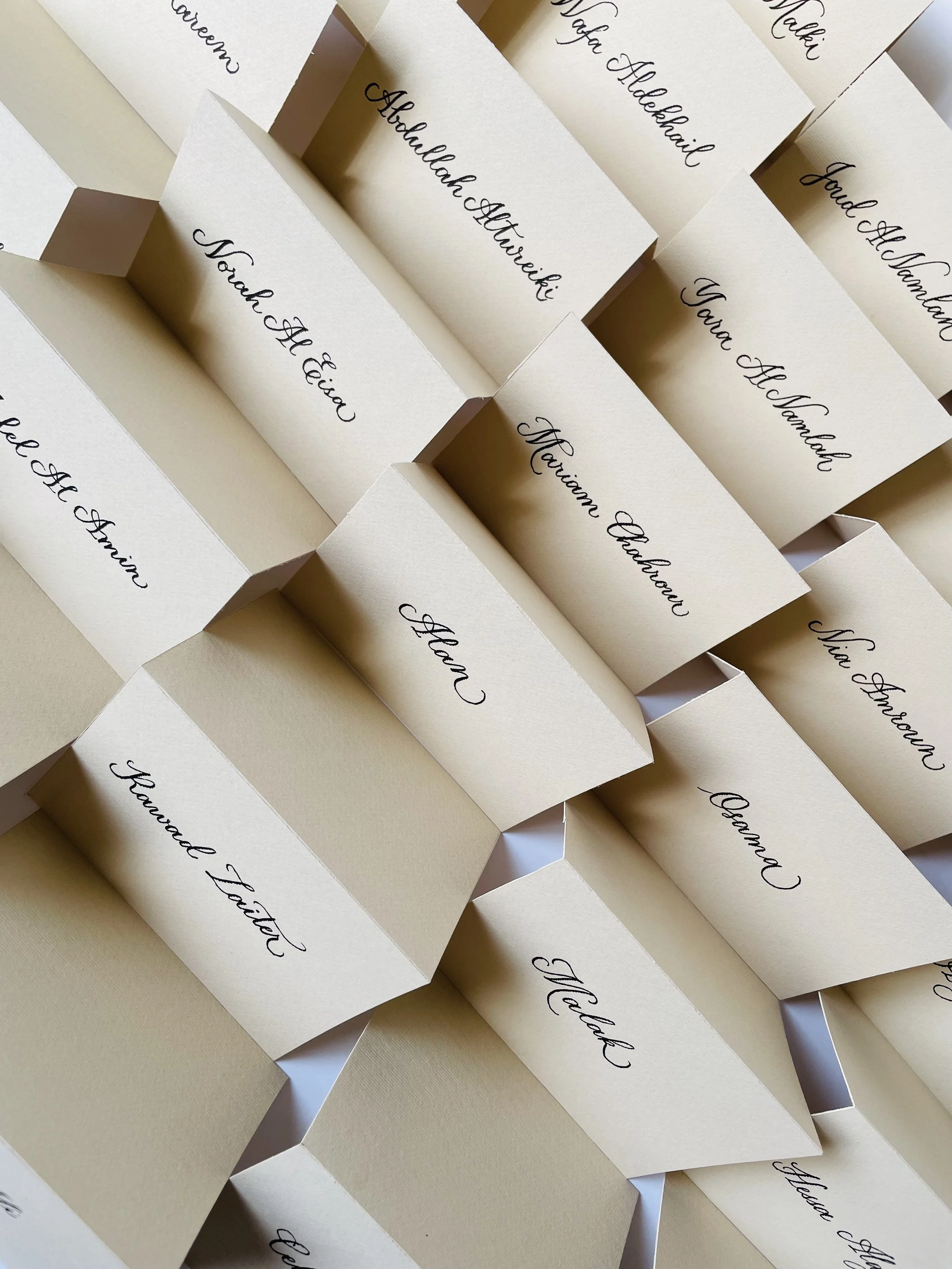 Multiple cream-colored place cards with black cursive calligraphy names such as Noah Alieva, Mariana Delgado, Yora Alomol, and Meli Gomez, arranged in a layered pattern.