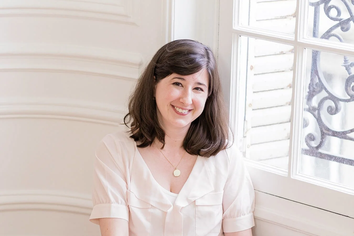 Michelle smiles into the camera. She is a white woman with dark brown hair. She is in a well-lit room with white walls, and leaning against a window.