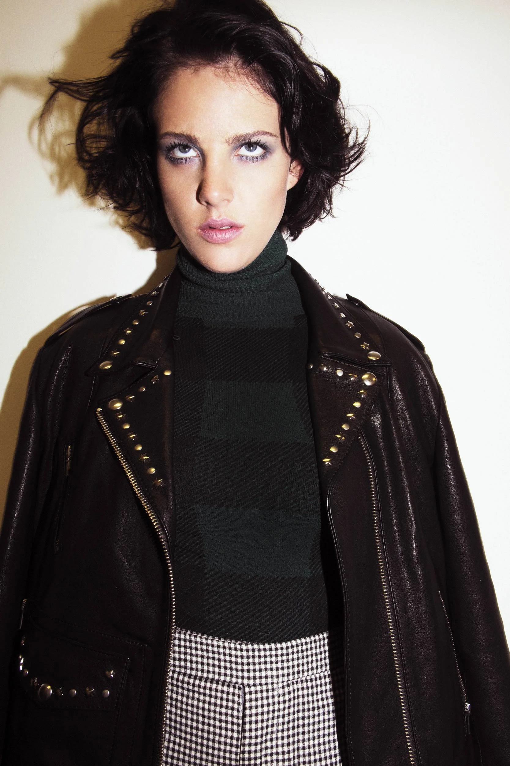 A woman in a dark green turtleneck and studded leather jacket stares upward with a skeptical expression, as if observing something strange above her. Her tousled hair and bold eye makeup add to the dramatic mood against a stark white background.
