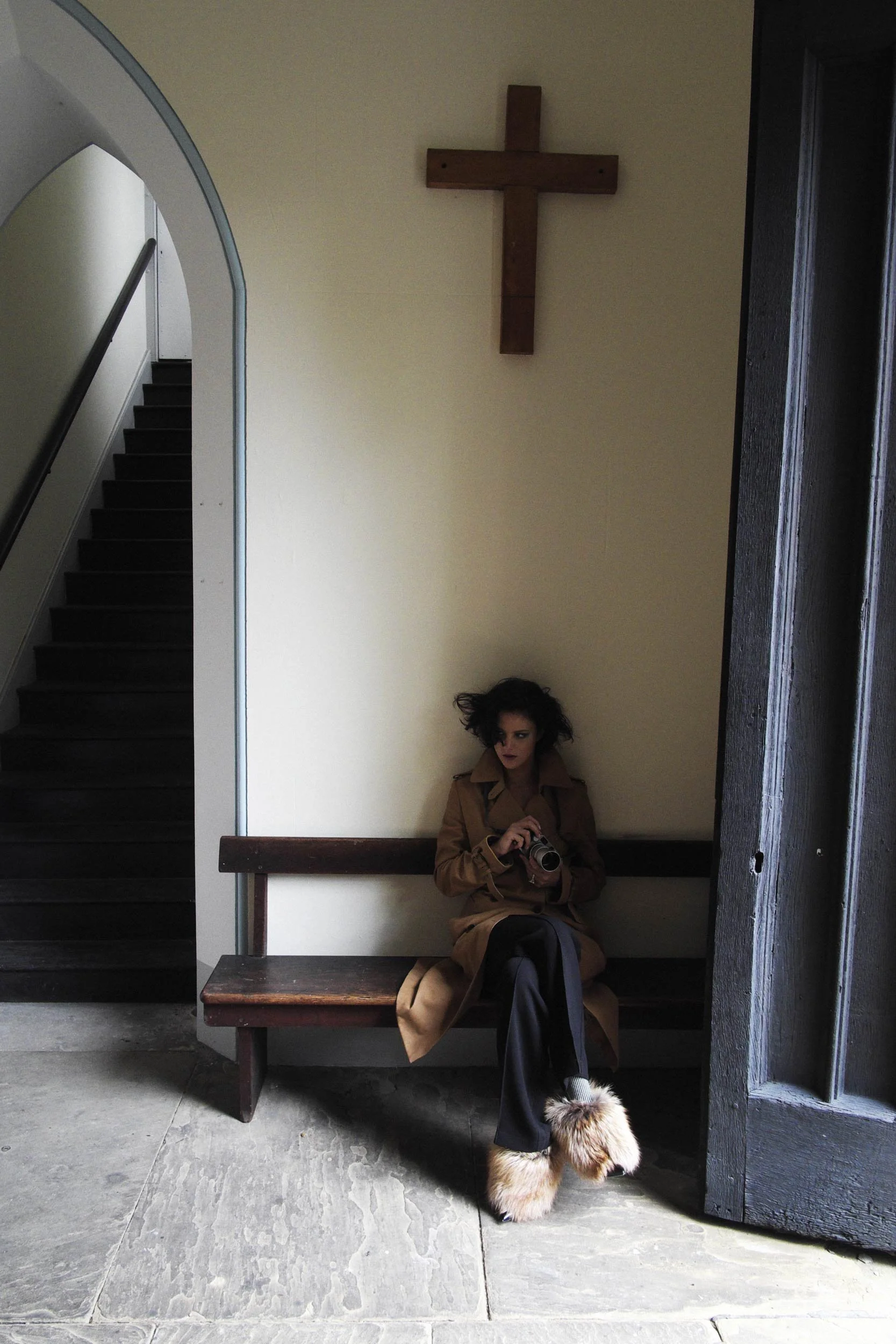 A woman sits on a wooden bench in a minimalist church interior, dressed in a tan trench coat and fur-trimmed boots, her dark curls tousled and moody lighting adding a cinematic effect.