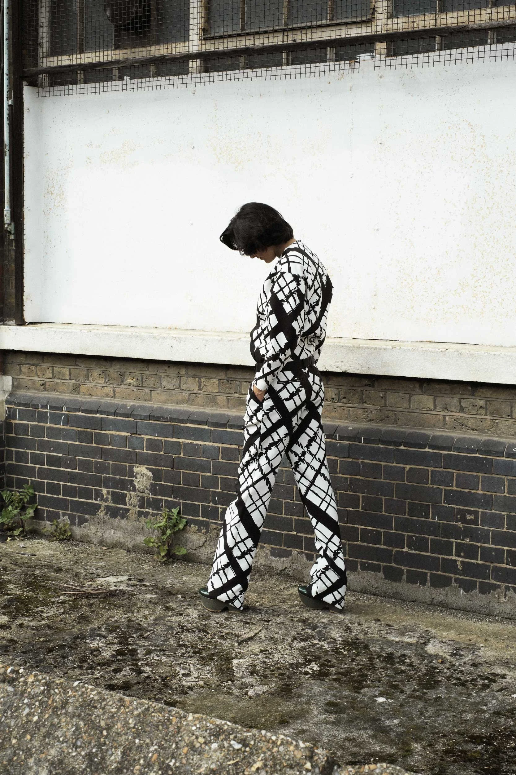 Fashion editorial featuring a model in a black and white printed ensemble, standing against a gritty brick wall with a wire fence in the background. The bold pattern contrasts with the urban textures.