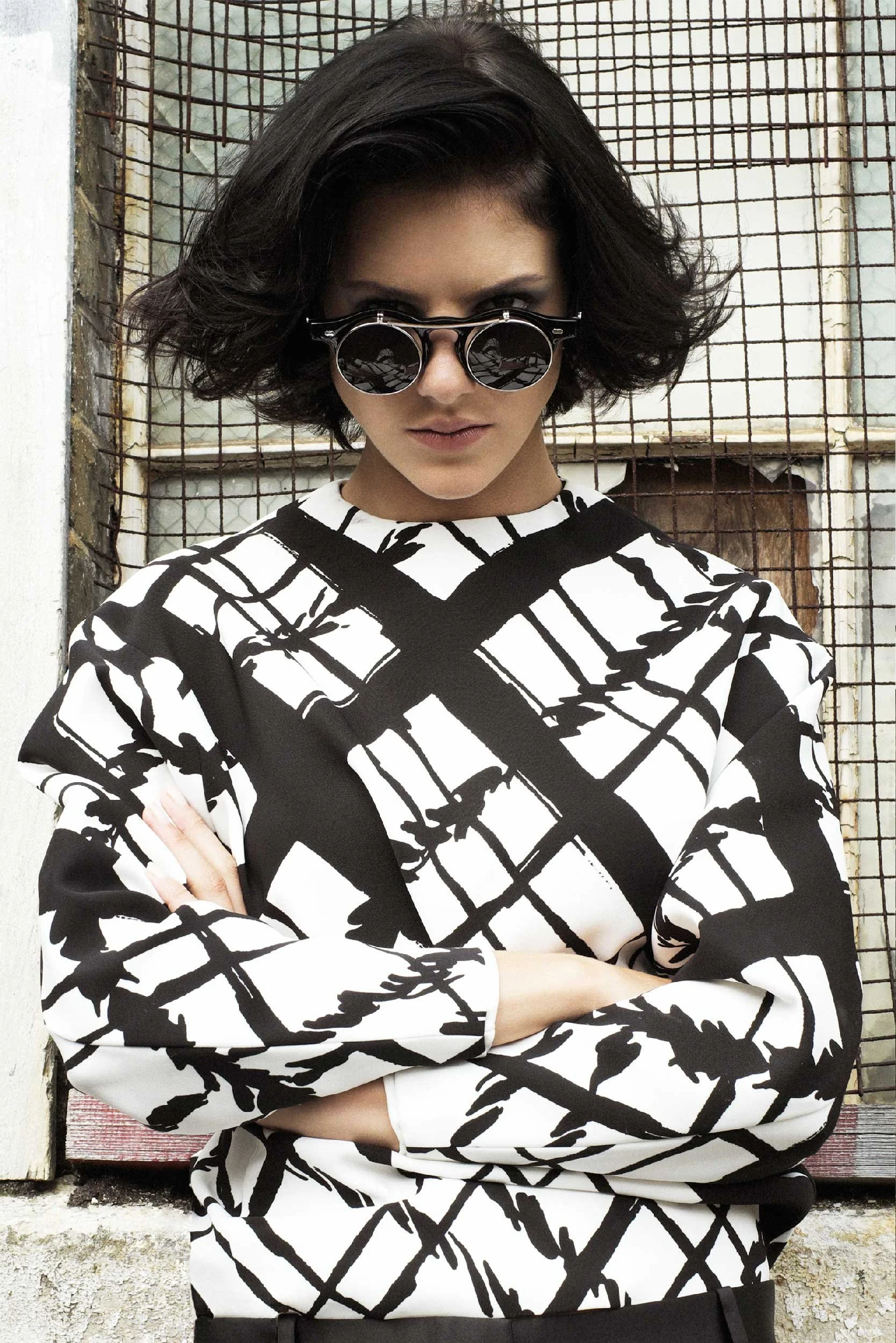 Editorial image of a model in an abstract black and white geometric patterned outfit, wearing round sunglasses with a strong, angular pose against an urban, industrial backdrop.