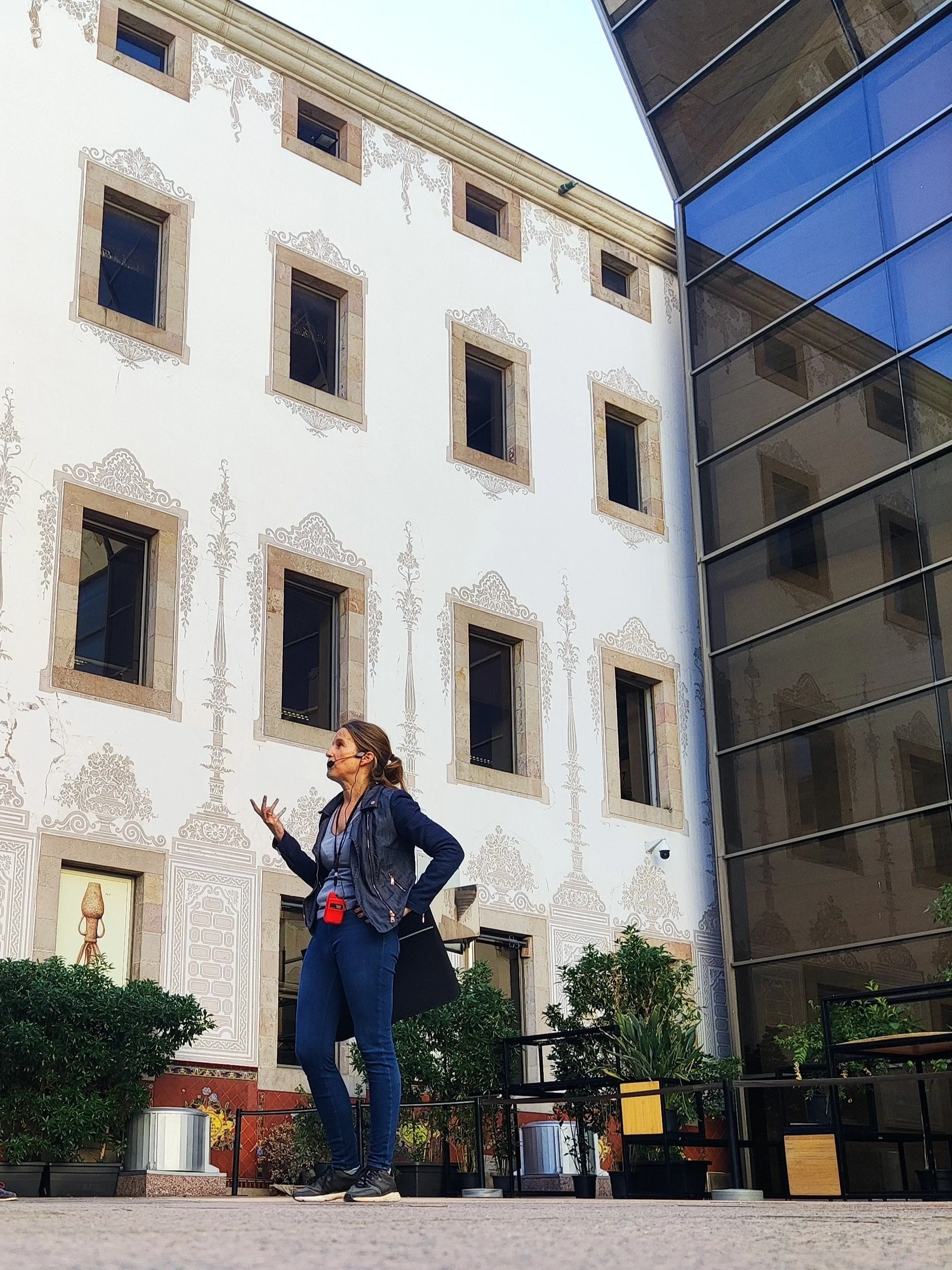 A woman speaking or giving a presentation outdoors in front of a white building with multiple small windows and decorative patterns, next to a modern glass building.