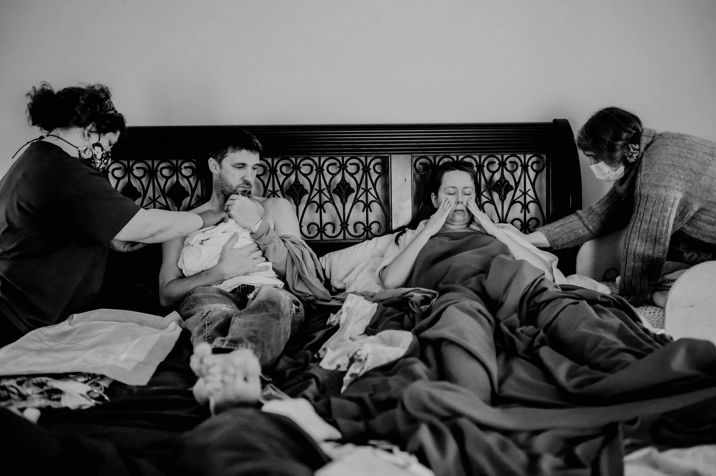 family sits together on bed at home after giving birth supported by doula and midwife