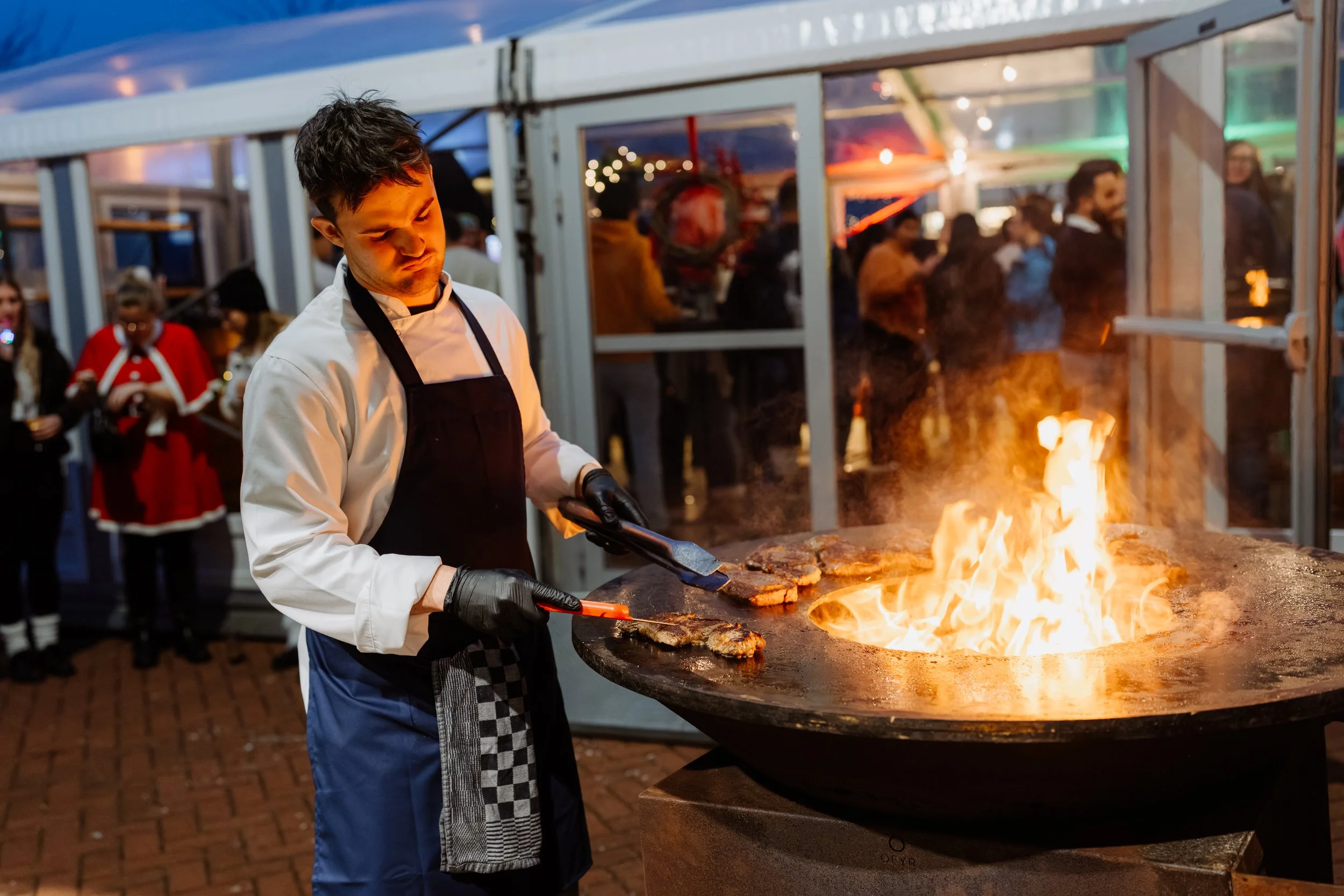 Kok bereidt steak op een grote gietijzeren barbecue met vlammen, in een feesttent met gasten op de achtergrond.
