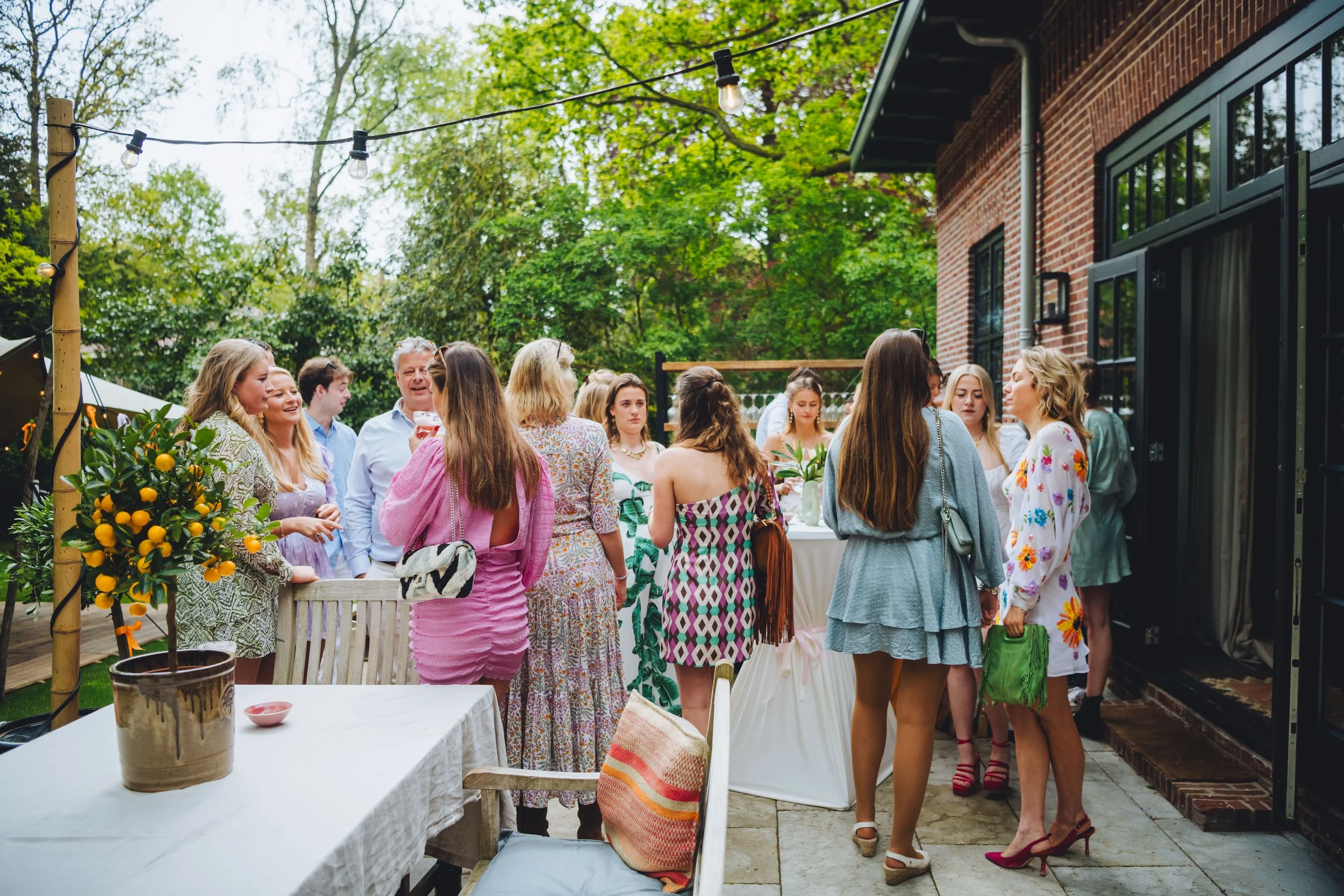 Mensen die samen in een tuinbijeenkomst staan, met bloemen, tafel, en zittende stoelen op de voorgrond.