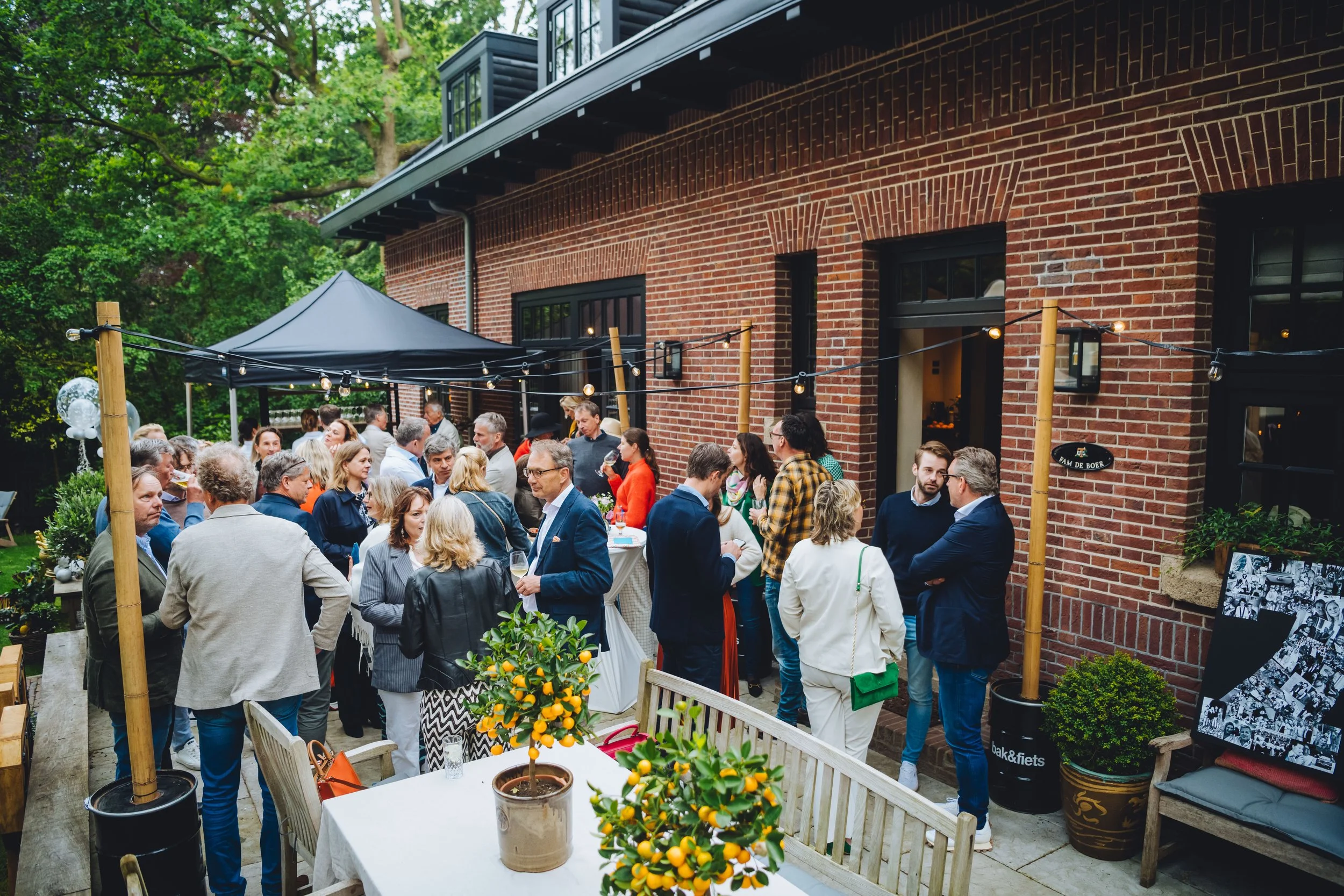 Een groepsfoto van een sociale bijeenkomst of feestje buiten bij een bakstenen huis, met mensen die praten en drinken onder een tent met stringverlichting.