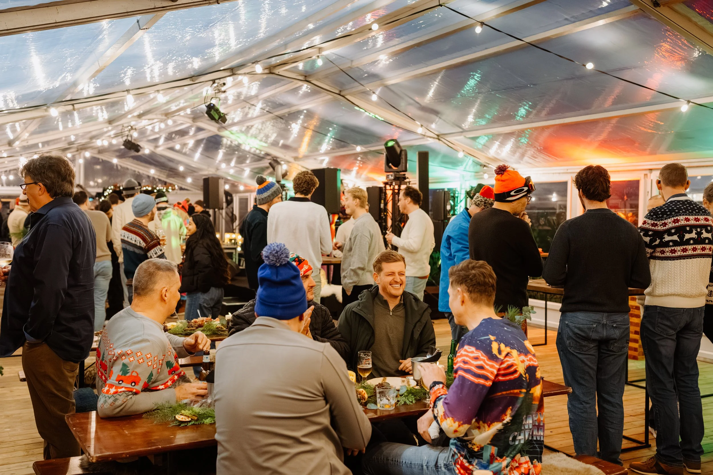 Gezellig kerstfeest met mensen in een feesttent, feestverlichting en decoraties, mensen zitten aan tafels en praten, sommige mensen dragen kersttruien en mutsen.