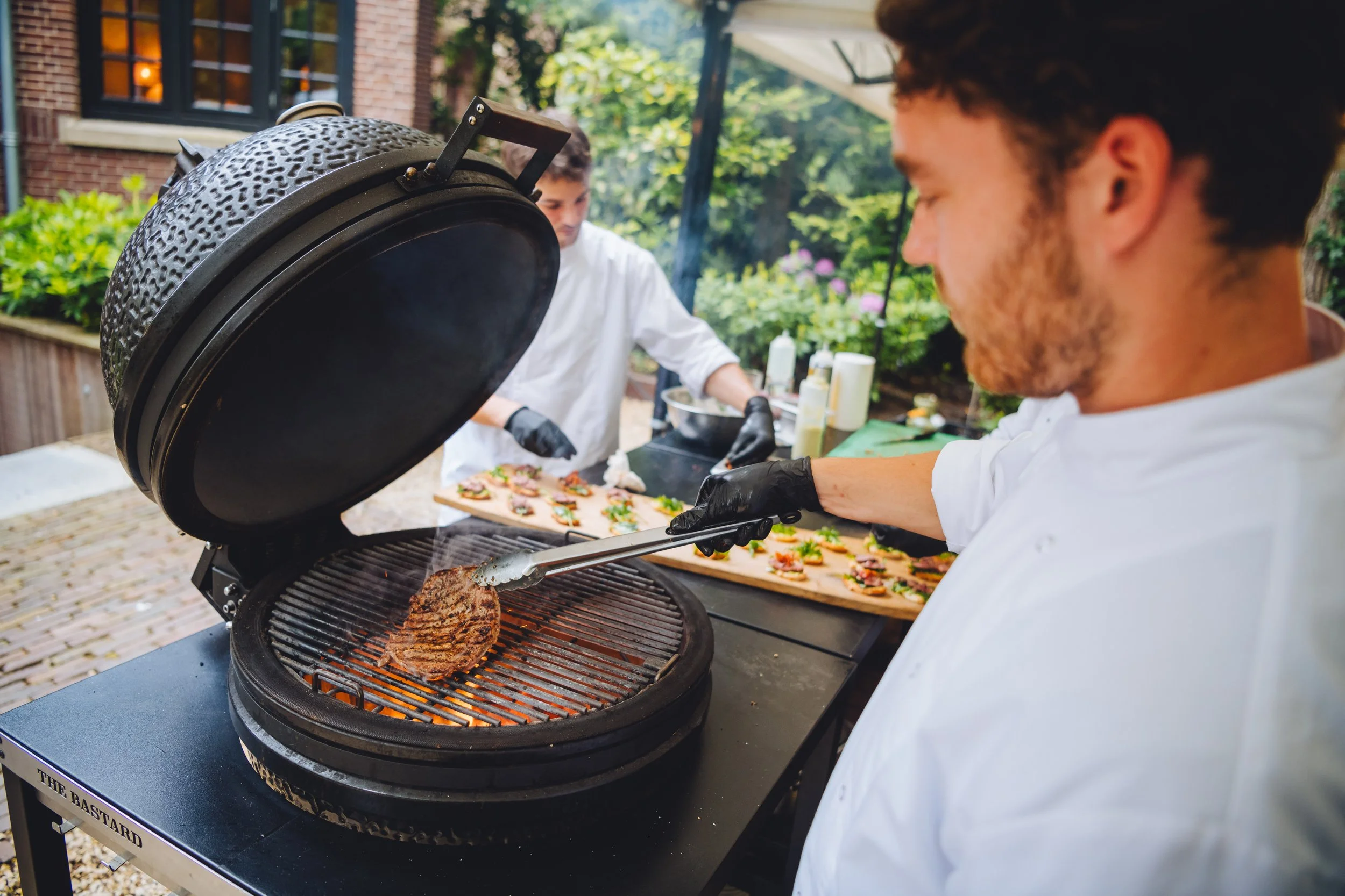 Twee mensen bereiden sandwiches en grillen een stuk vlees op een barbecue in een tuin.