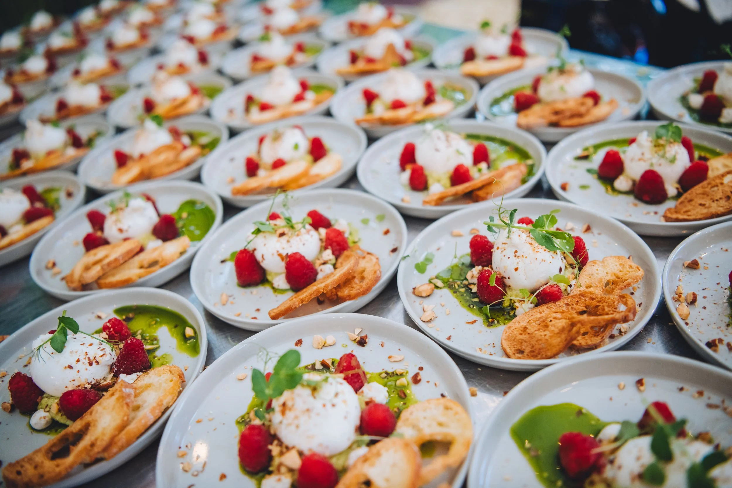 Een aantal borden met een gerecht bestaande uit een bol van witte kaas, frambozen, groene saus, een krokant stuk brood en groene kruiden, mooi gepresenteerd.