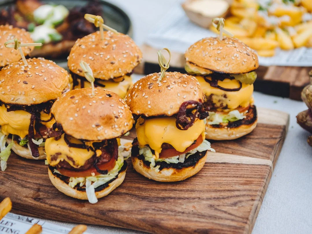 Mini cheeseburgers met sesambroodjes, kaas, sla, tomaat en augurk, op een houten plank.