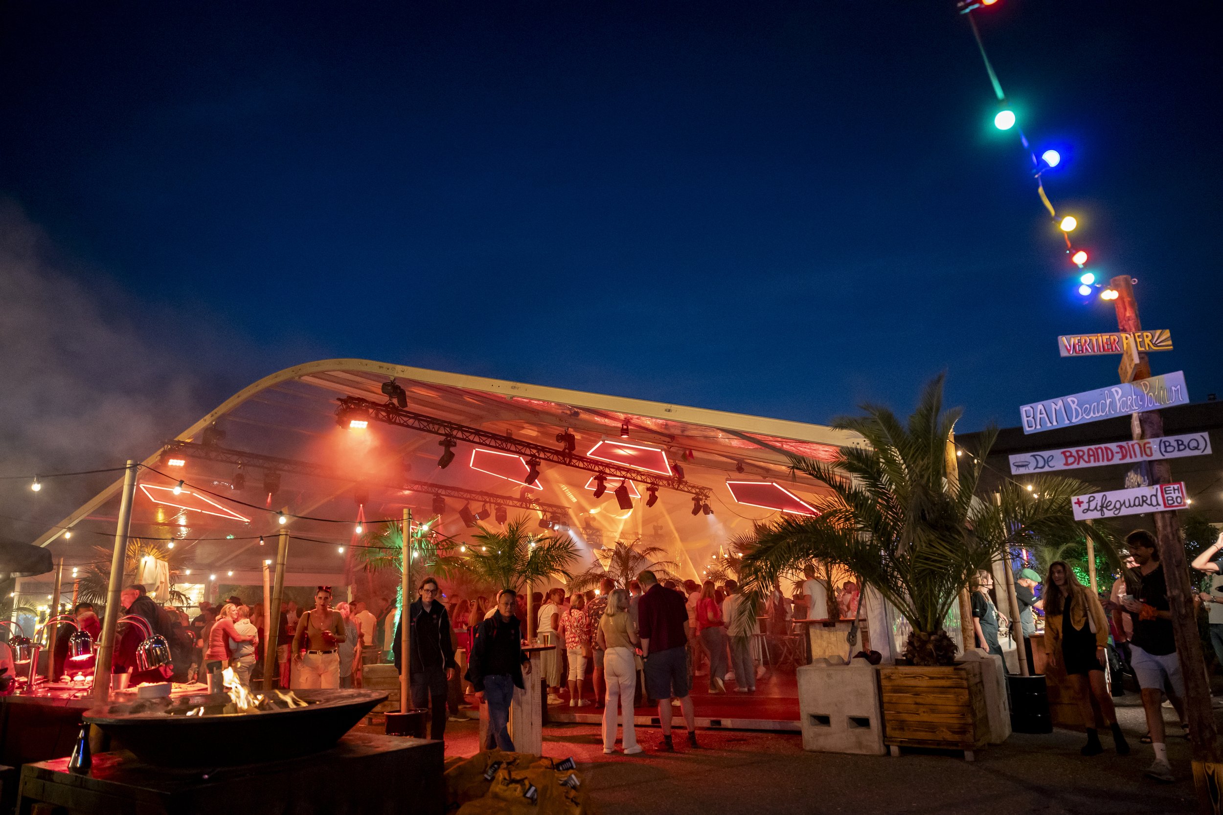 Een feesttent bij zonsondergang met kleurrijke verlichting, mensen die binnen en buiten staan, en sierlijke palmbomen met kleurrijke borden met teksten.