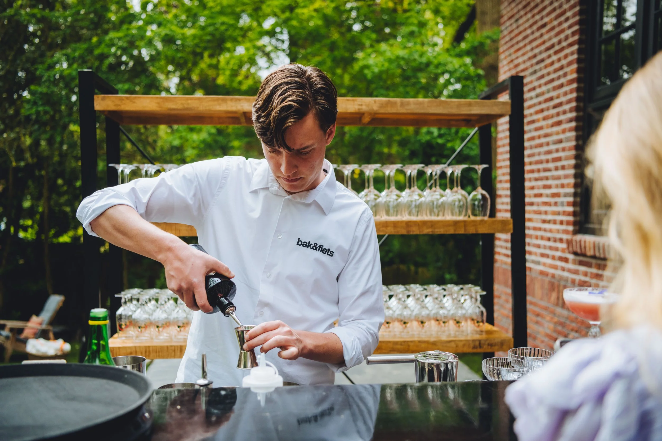 Barman bereidt een cocktail met een shaker, in een buitenruimte met flesjes en glazen op de achtergrond.