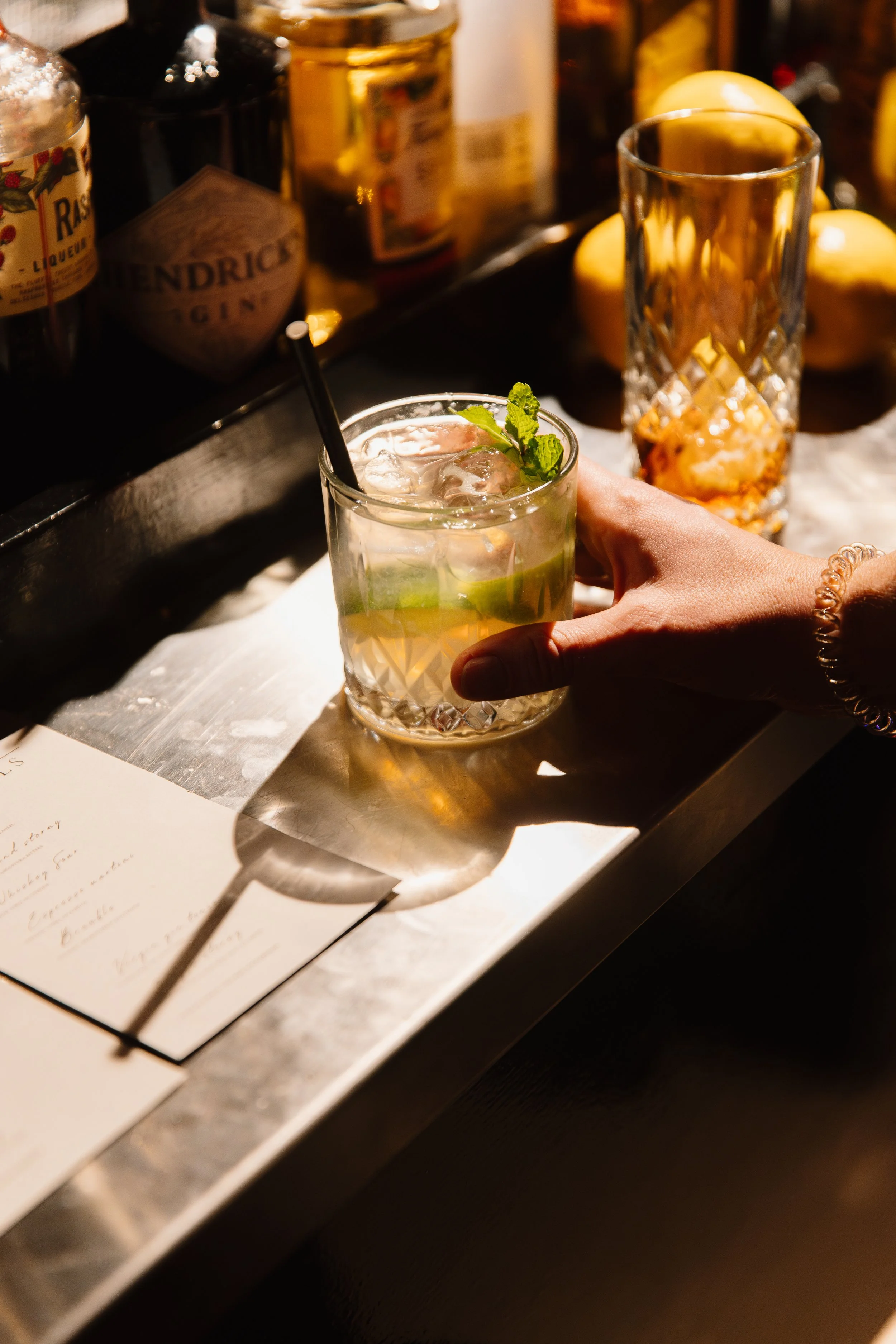 Een hand vasthoudt een glas met een cocktail, versierd met munt en limoen, op een bar. Op de achtergrond zijn verschillende flesjes en een lege glas.