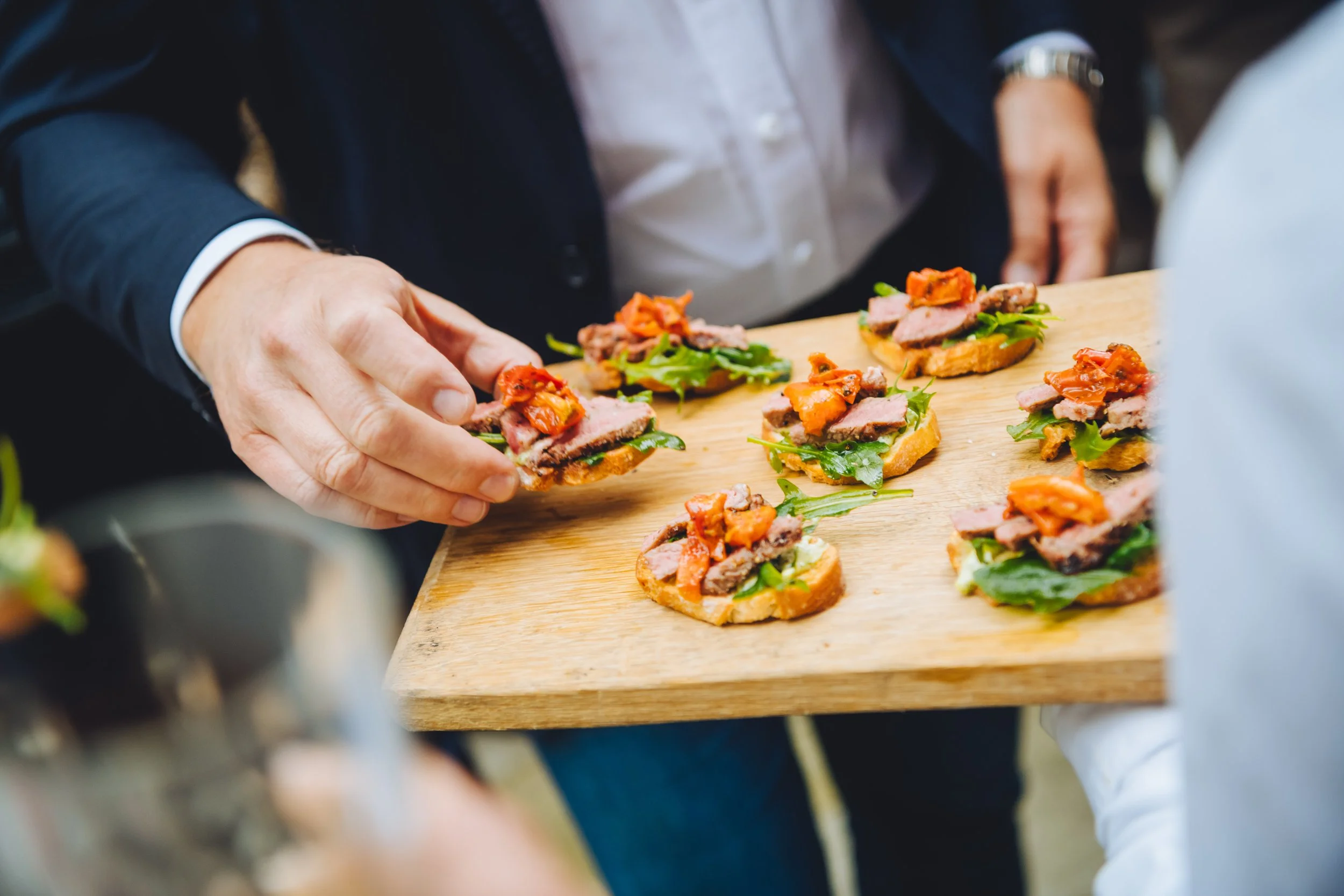 Een persoon serveert kleine hapjes met vlees, groene bladeren en pittige saus op een houten plank.