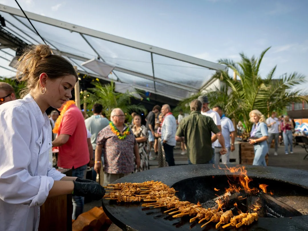 Vrouw bereidt gegrilde spiesen met vlees op een grote grill tijdens een buitenfeest met een groep mensen onder een tent en omringd door palmbomen.