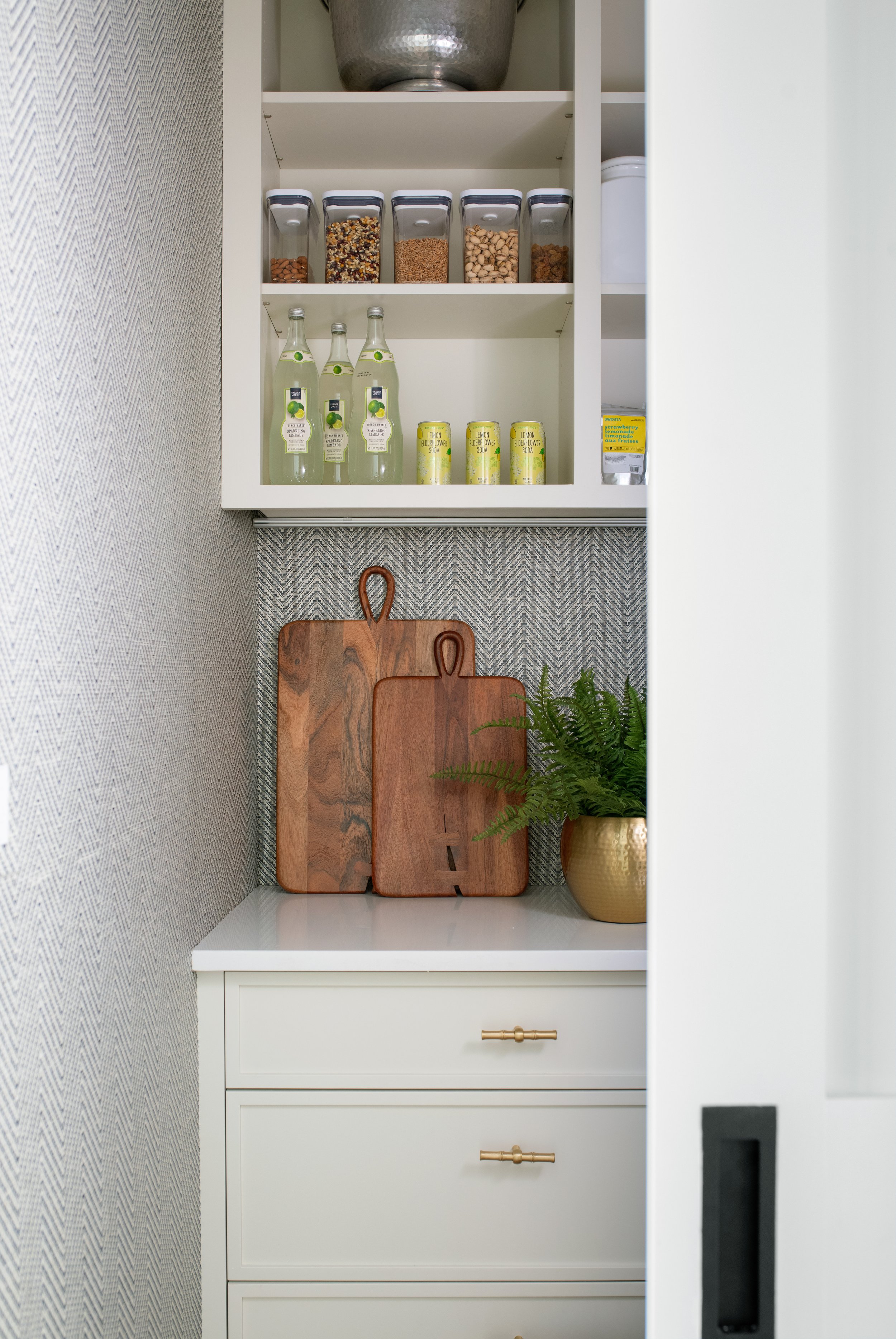 A coastal kitchen pantry with herringbone textured wallpaper, brass accents, and white cabinetry.