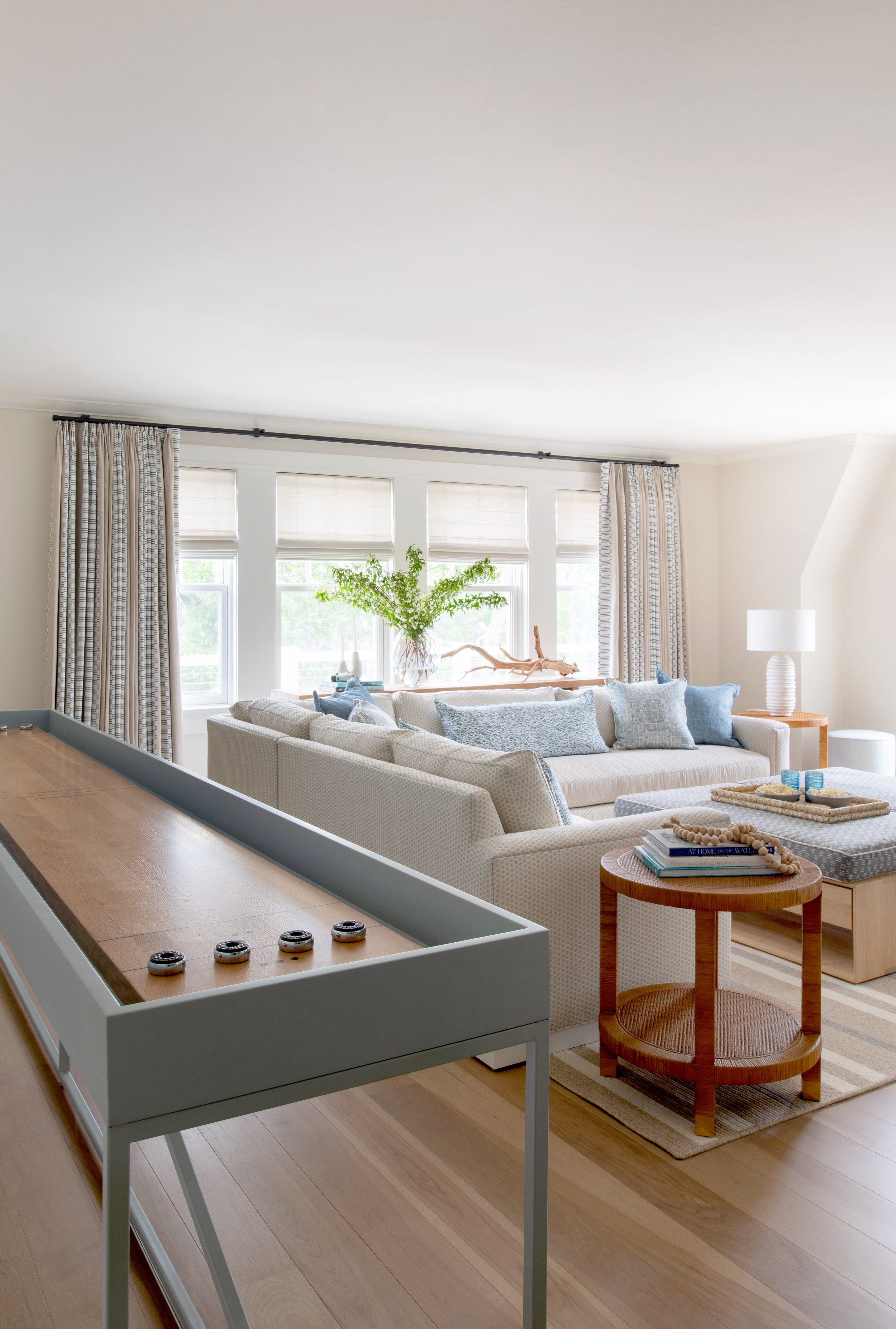 Coastal sitting room with a simple blue shuffle-board table, wide plank wood floors, and floor to ceiling drapery.