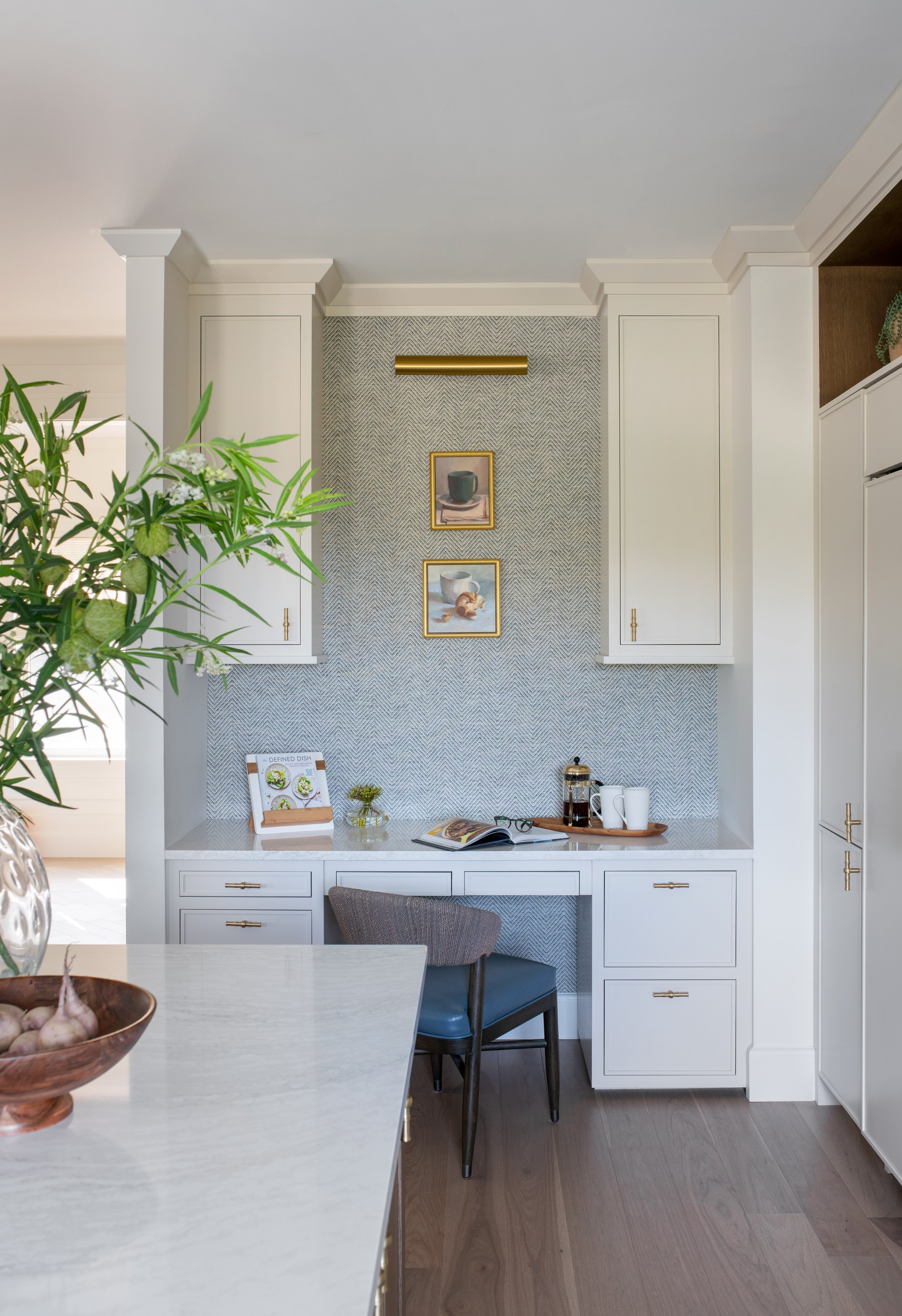 An all white kitchen with natural accents, a built-in desk, brass hardware, a picture light, and accent wallpaper. Coastal Kitchen Scheme.