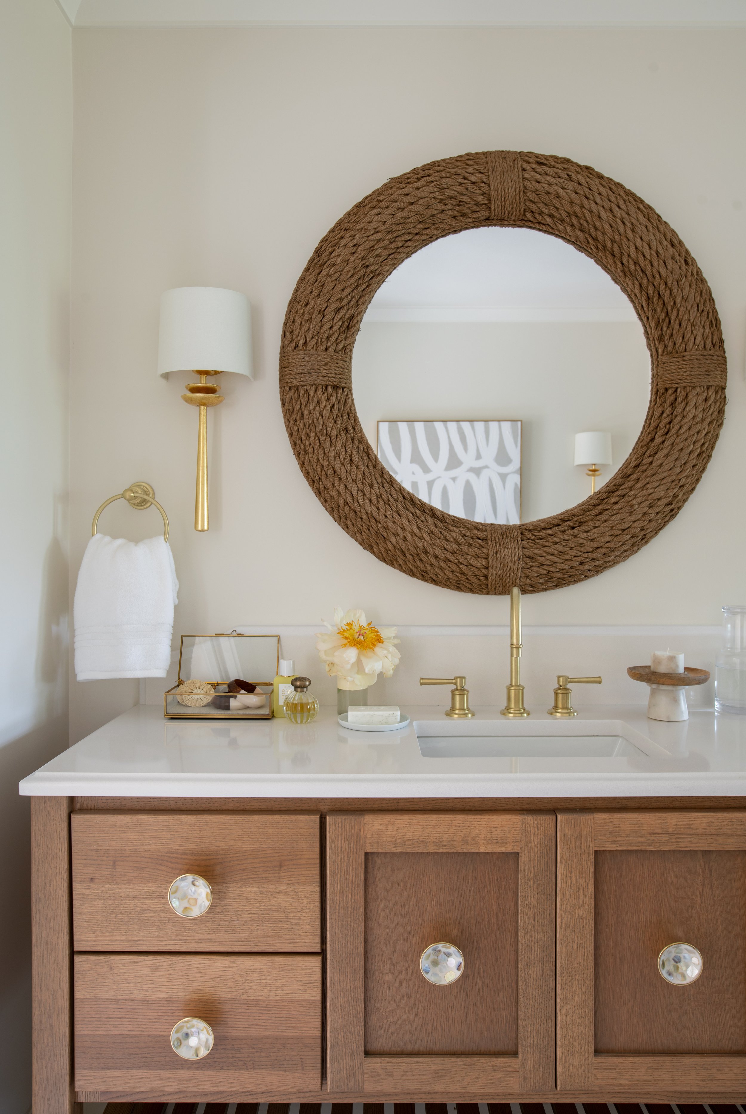 natural wood vanity with a white countertop, brass faucet, brass sconces, and fun cabinet hardware, with a woven coastal mirror.
