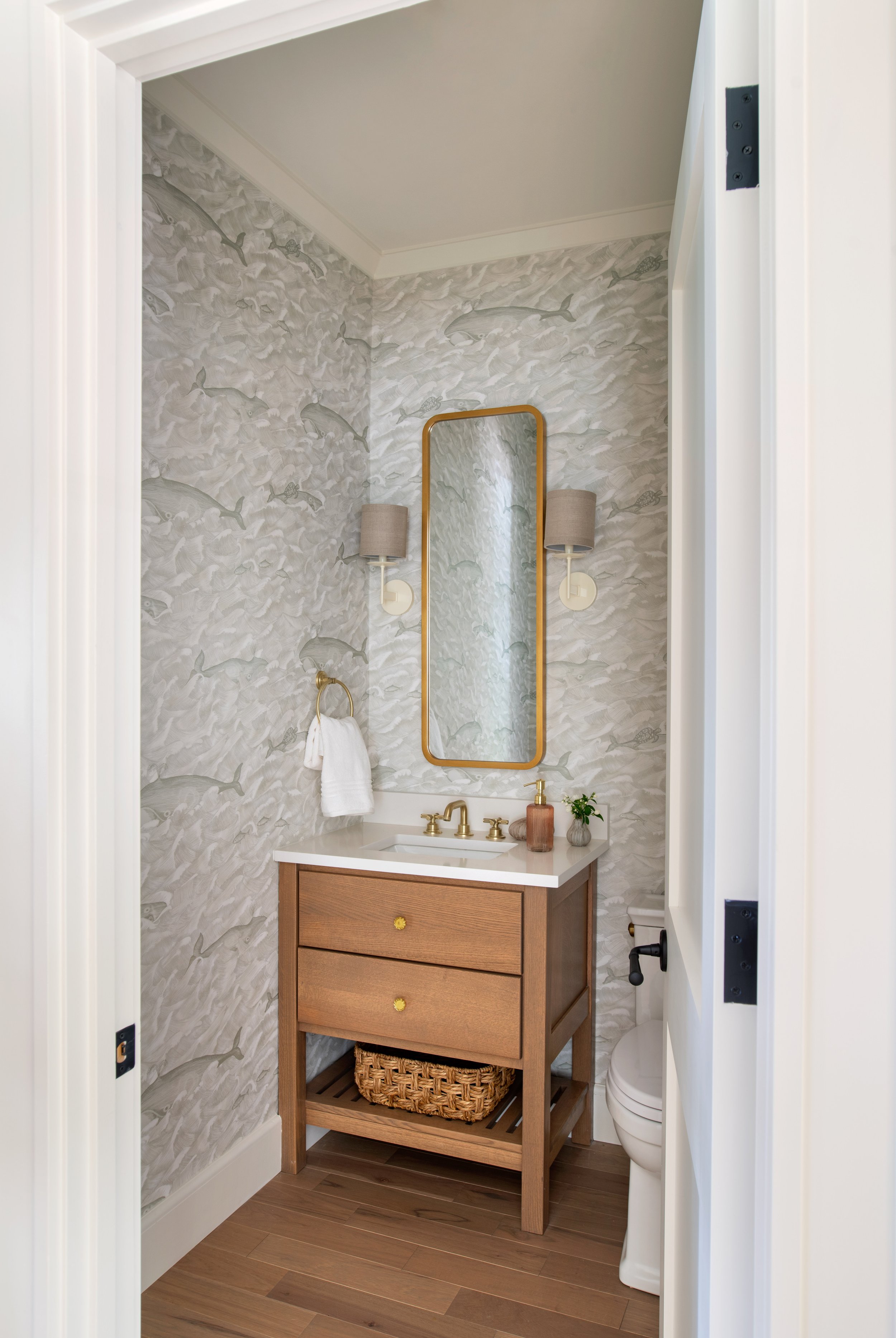 Coastal powder room with a natural wood vanity, grey whale-themed wallpaper, off-white wall sconces, and a large brass mirror.