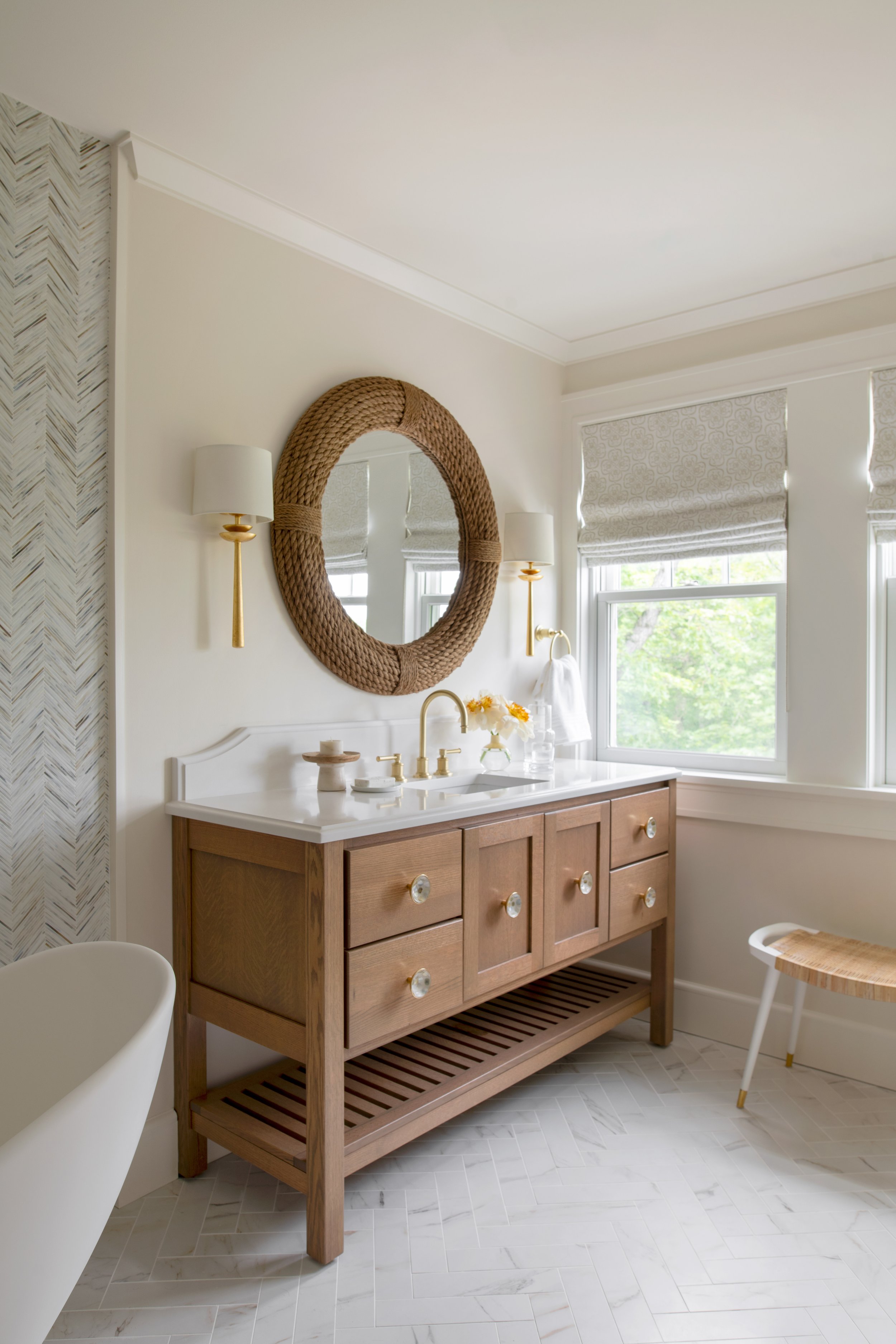Coastal design style master bathroom with natural wood vanities and white countertops, herringbone wall tile, and brass hardware and wall sconces.