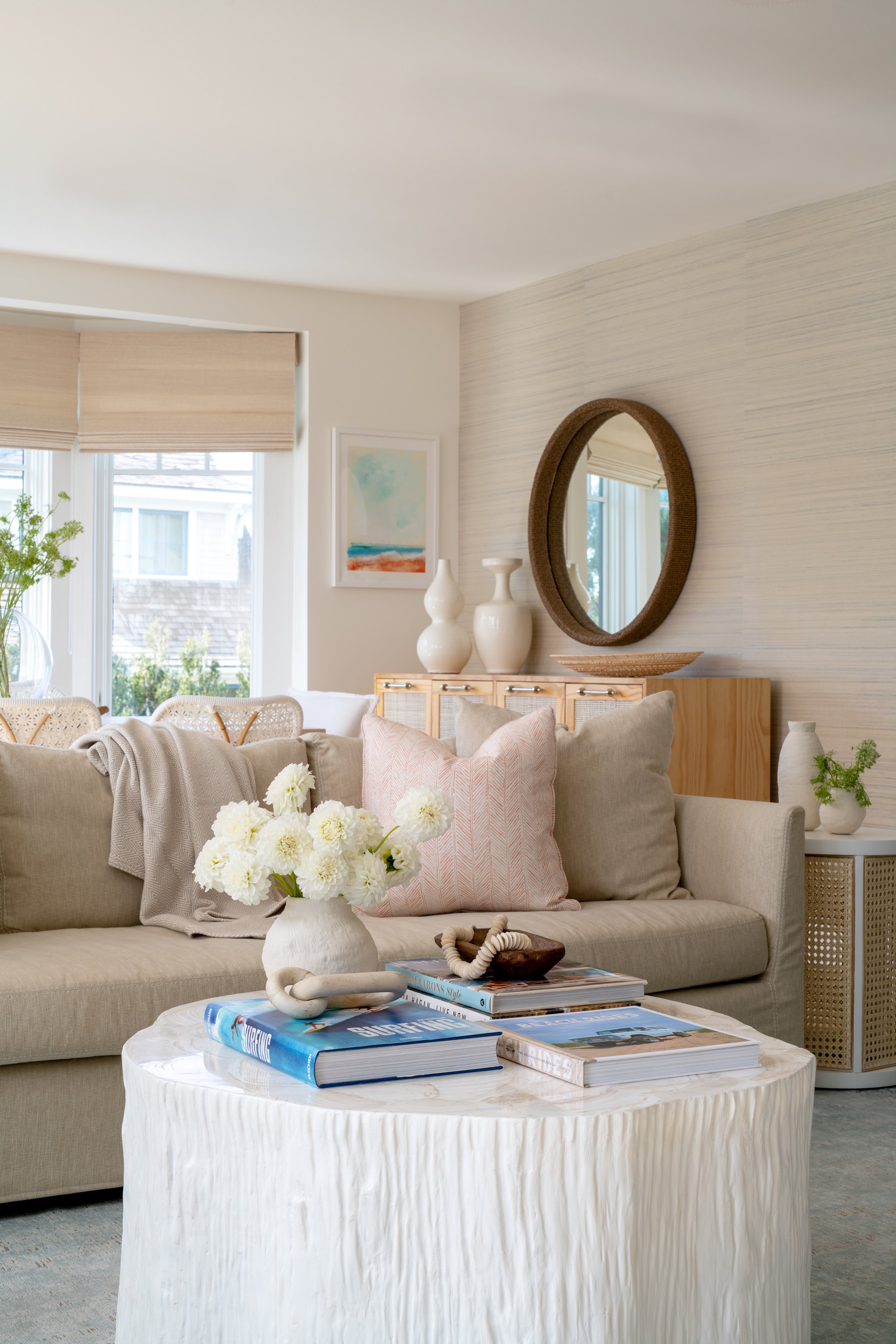 Coastal living room with blue and pink accents. White, textured coffee table. Coastal coffee table books. Beige grasscloth wall covering. Coastal wall covering. Coastal window coverings.