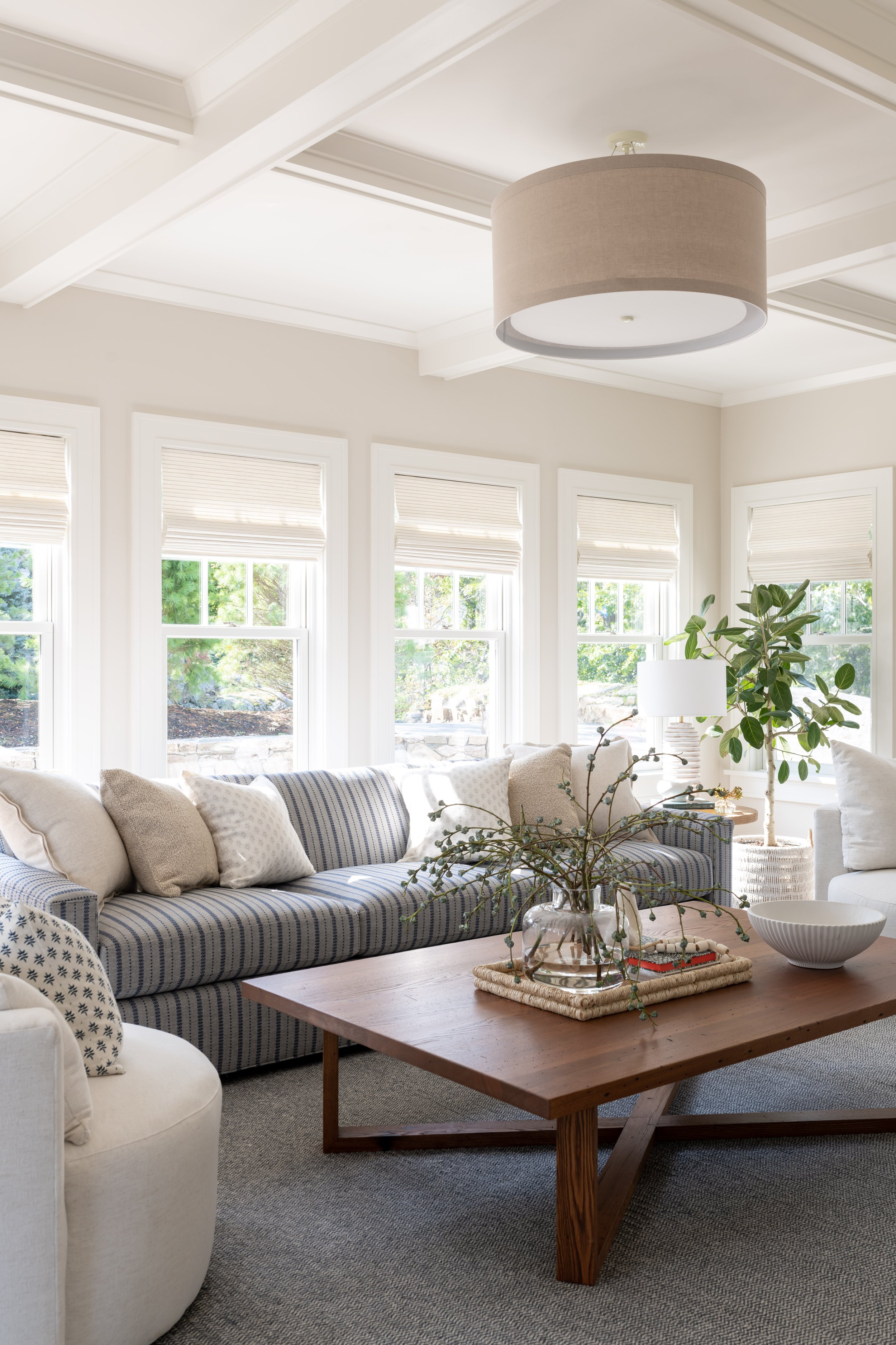 A bright, cozy sitting room with a large natural wood coffee table, a striped sofa, and oversized swivel chairs. Coffered Ceilings. Coastal Style.