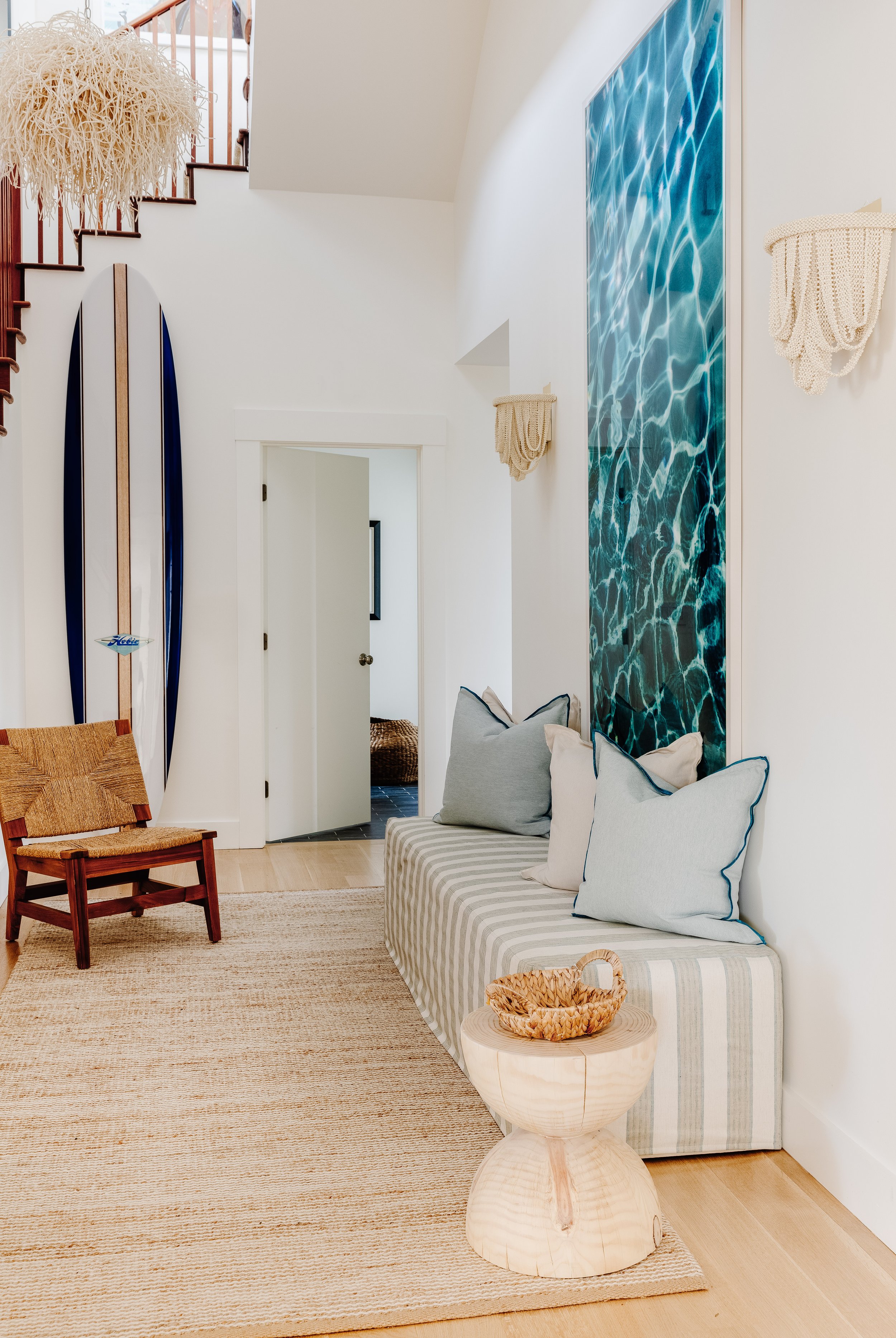 Coastal entry hall with a striped accent bench, wooden side table, woven accent chair, and beaded accent lighting. Beaded wall sconces in a coastal hallway. Blue accent pillows.