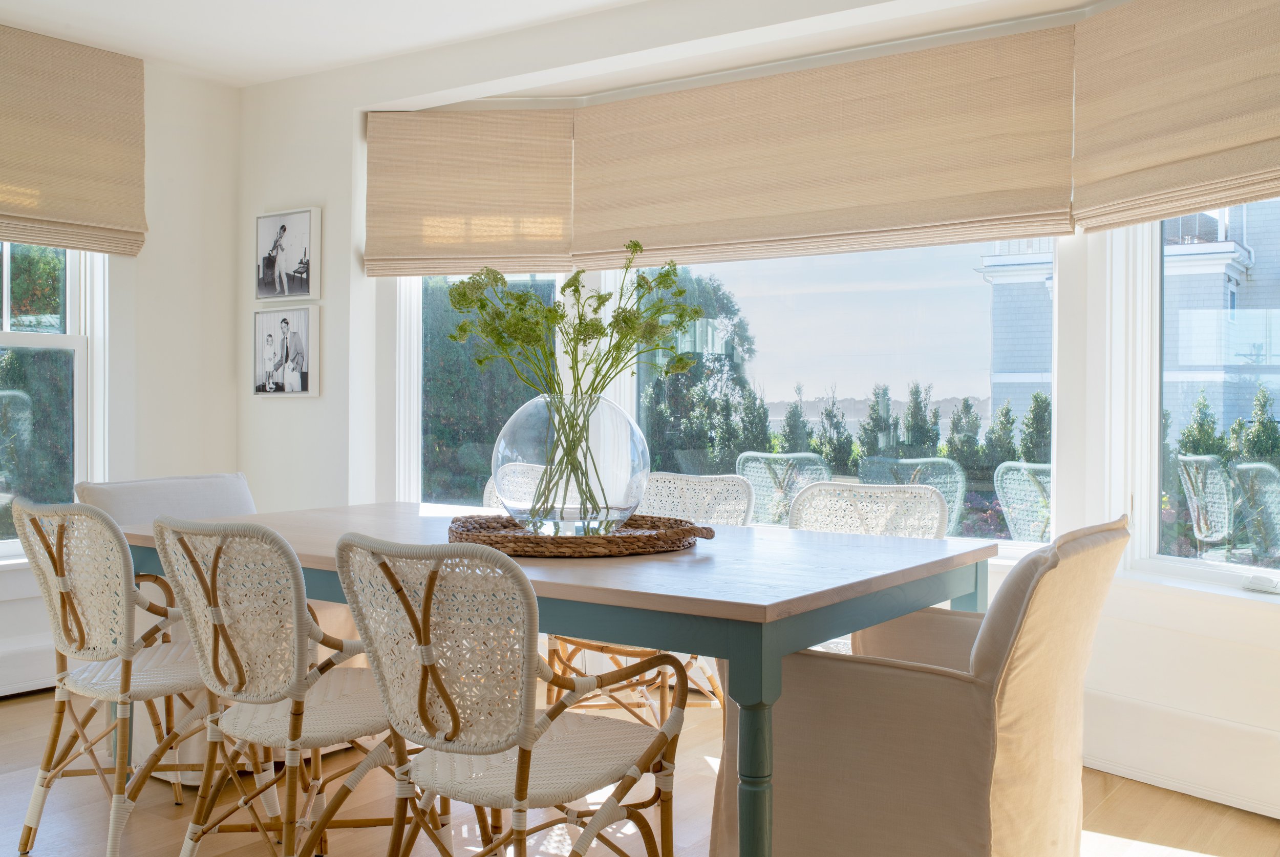 Large coastal dining area with large windows accented by woven wood roman shades. Large coastal dining table with a blue base and white top. White bistro-style woven chairs and wood accents. Coastal dining table decor. North Shore Massachusetts Coast