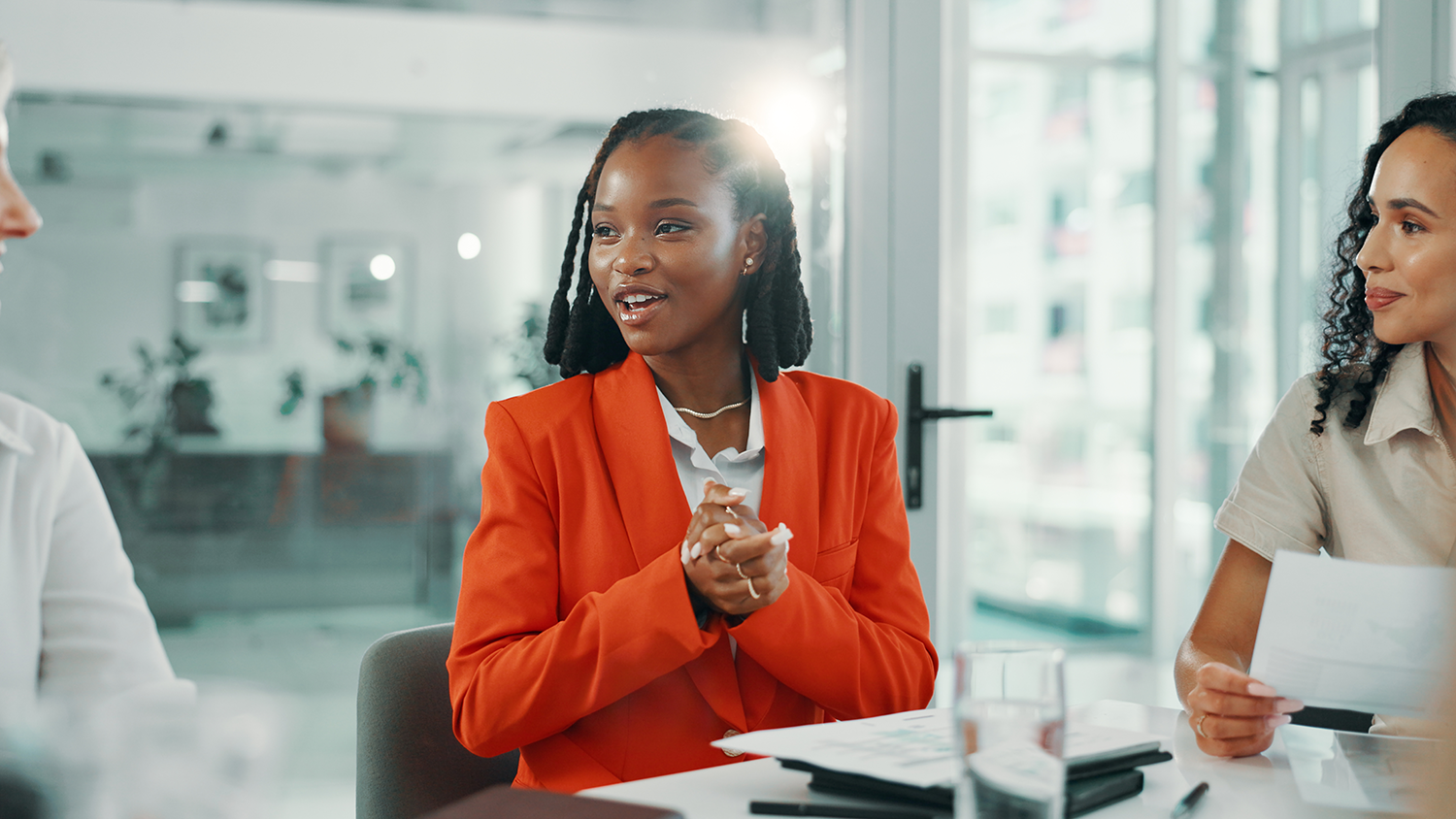 Businesswoman in an orange blazer talking during a meeting with two women, one on her left and one on her right, in a modern office with large windows.