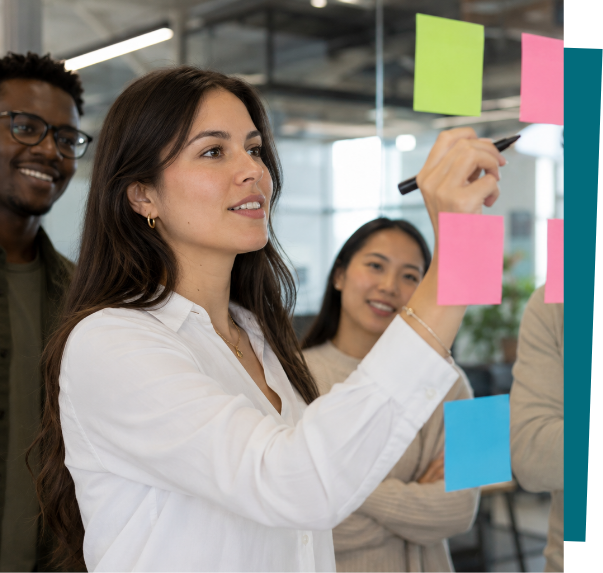 A woman writing on colorful sticky notes on glass in a modern office with two smiling colleagues observing.