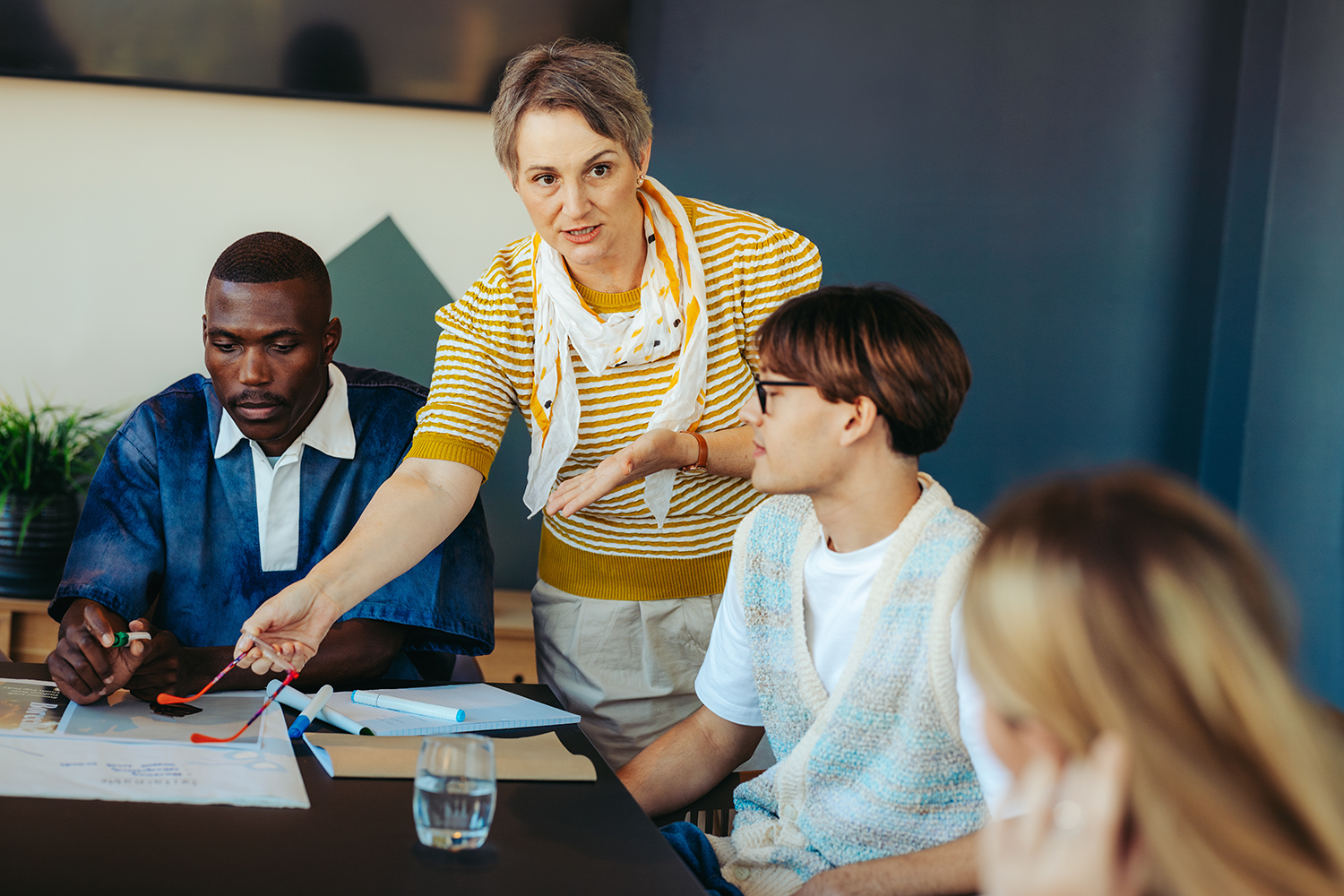 A woman with short gray hair wearing a yellow and white striped shirt and white scarf is talking to three young men during a meeting. The men are seated around a black table with papers and markers, with a glass of water visible on the table.