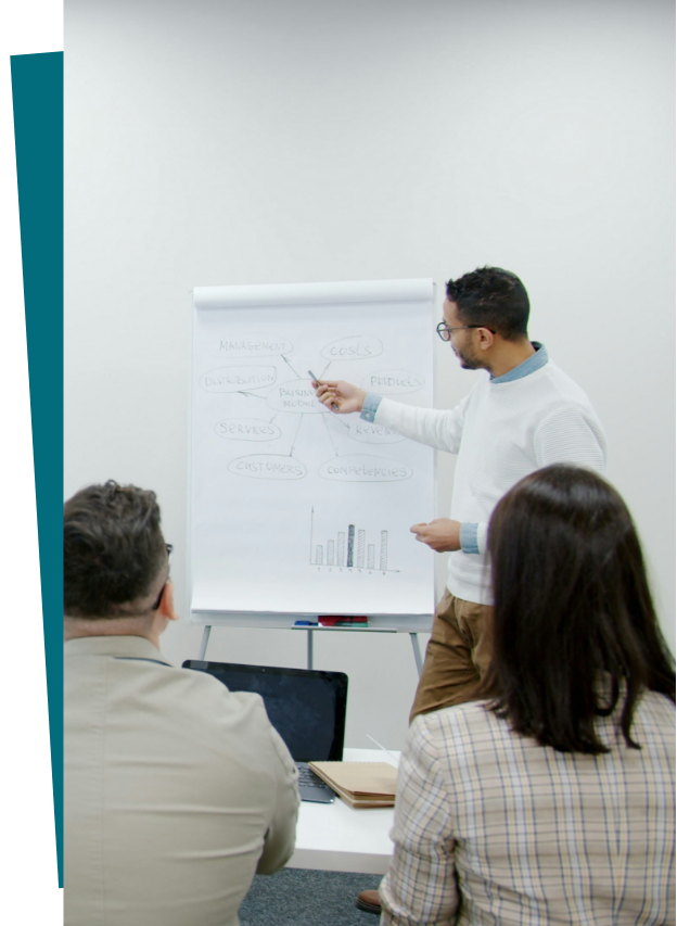 A man presenting a diagram on a whiteboard during a business meeting with two seated colleagues taking notes.