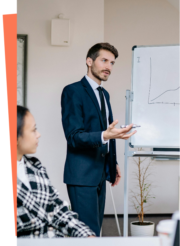 A man in a navy suit giving a presentation using a whiteboard, with a woman in a black and white patterned coat listening in a conference room.