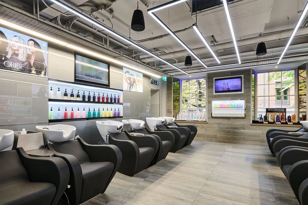 Interior of a modern hair salon with black and white chairs at wash stations, wall-mounted screens, and large windows showing trees outside.