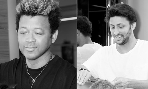 Two men sitting at a table, one with curly hair looking down and the other with straight hair smiling and looking at something on the table