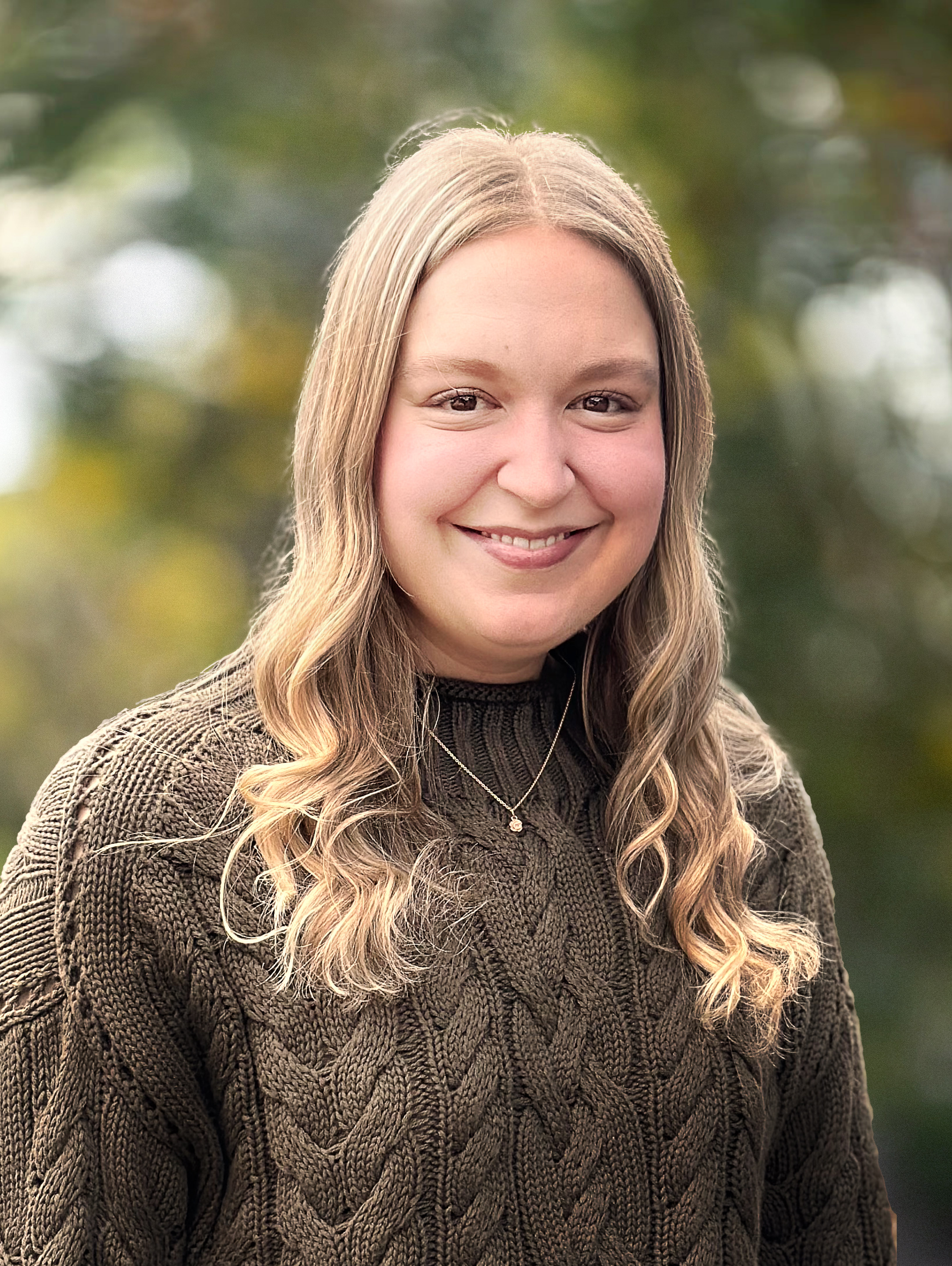 A woman with long blonde hair smiling outdoors with blurred greenery in the background.