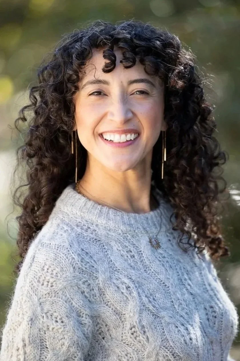 A woman with curly dark hair and gold earrings smiling in front of a blurred background.