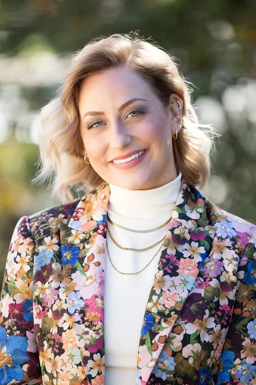 A woman with blonde, wavy hair and light makeup, smiling, wearing layered necklaces and earrings, dressed in a floral blazer, standing outdoors with blurred green foliage in the background.