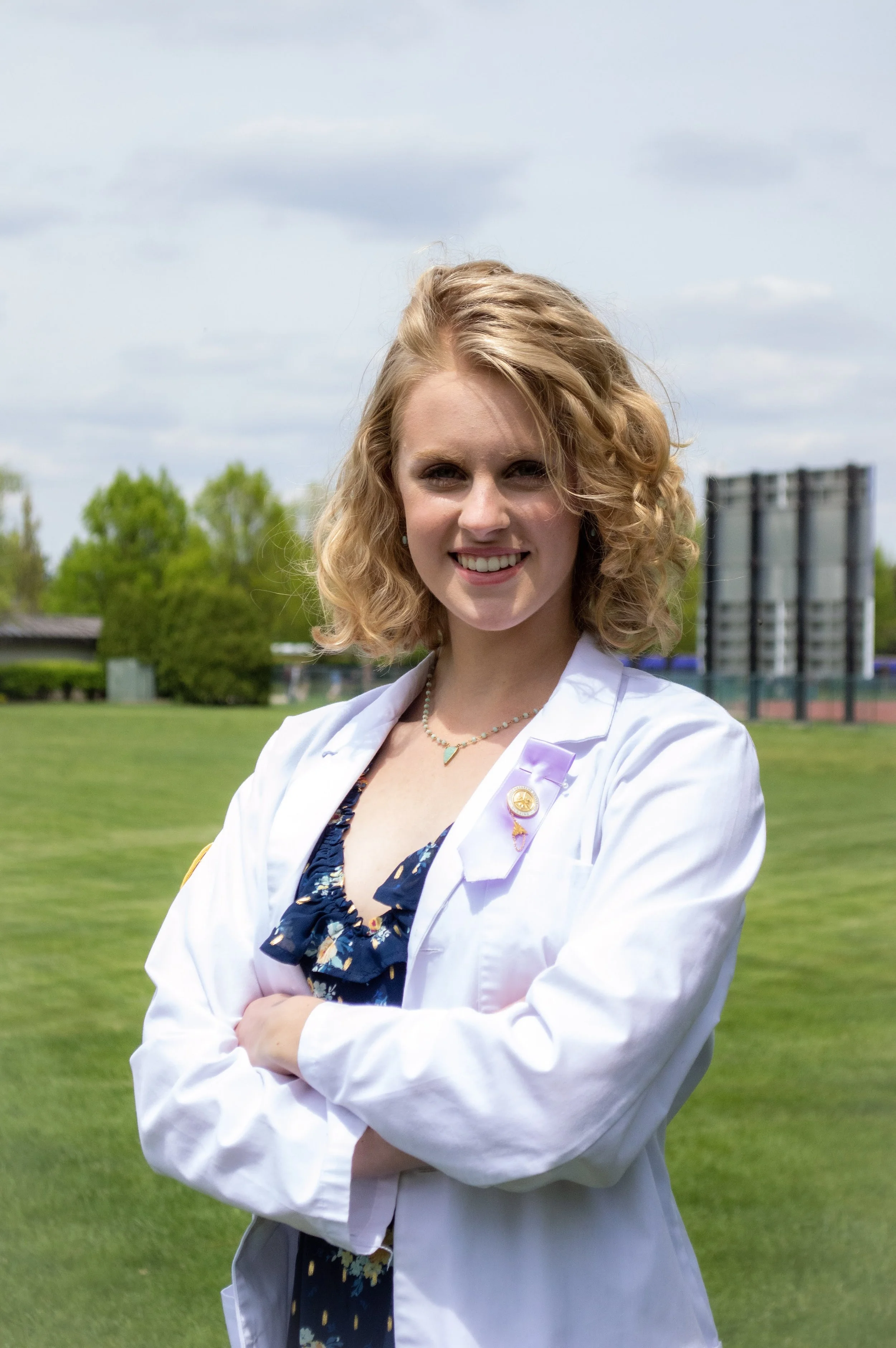Person with curly blonde hair in a white lab coat standing on a grassy field, smiling.