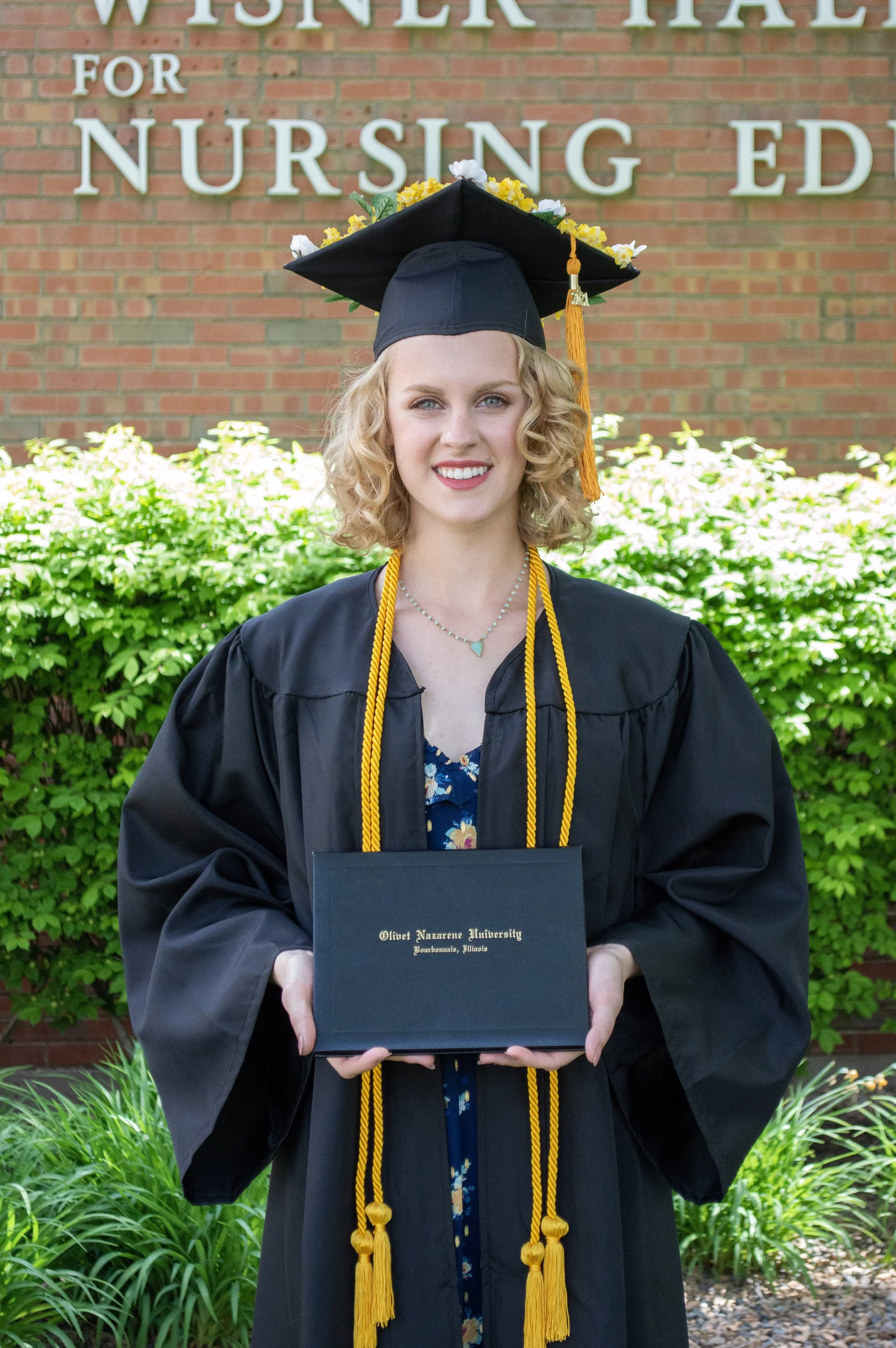 Graduate in cap and gown holding diploma in front of a building labeled "Wisner Hall for Nursing Education."
