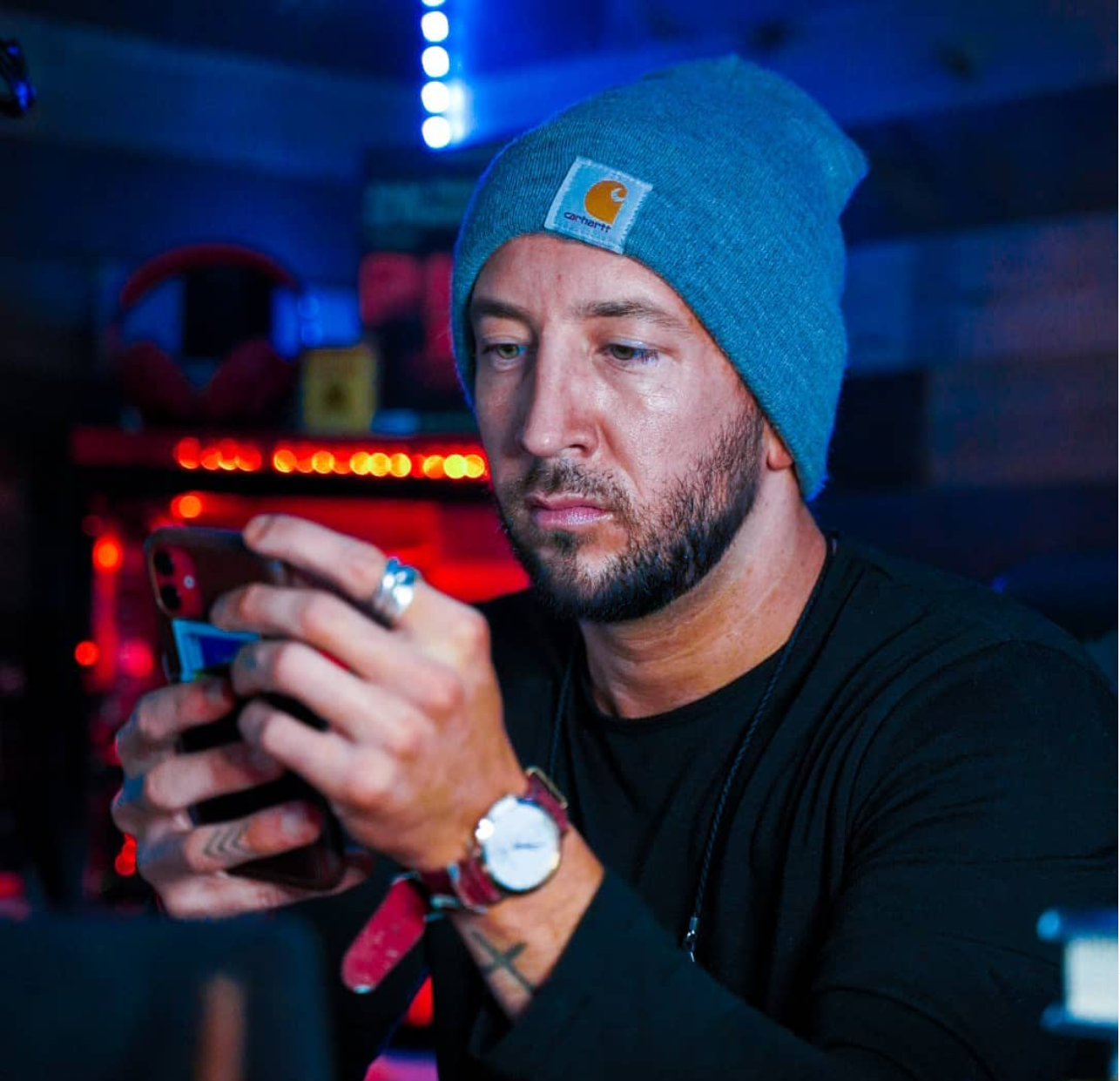 Man in blue beanie and black shirt using a smartphone in a dimly lit room with red and blue lights.