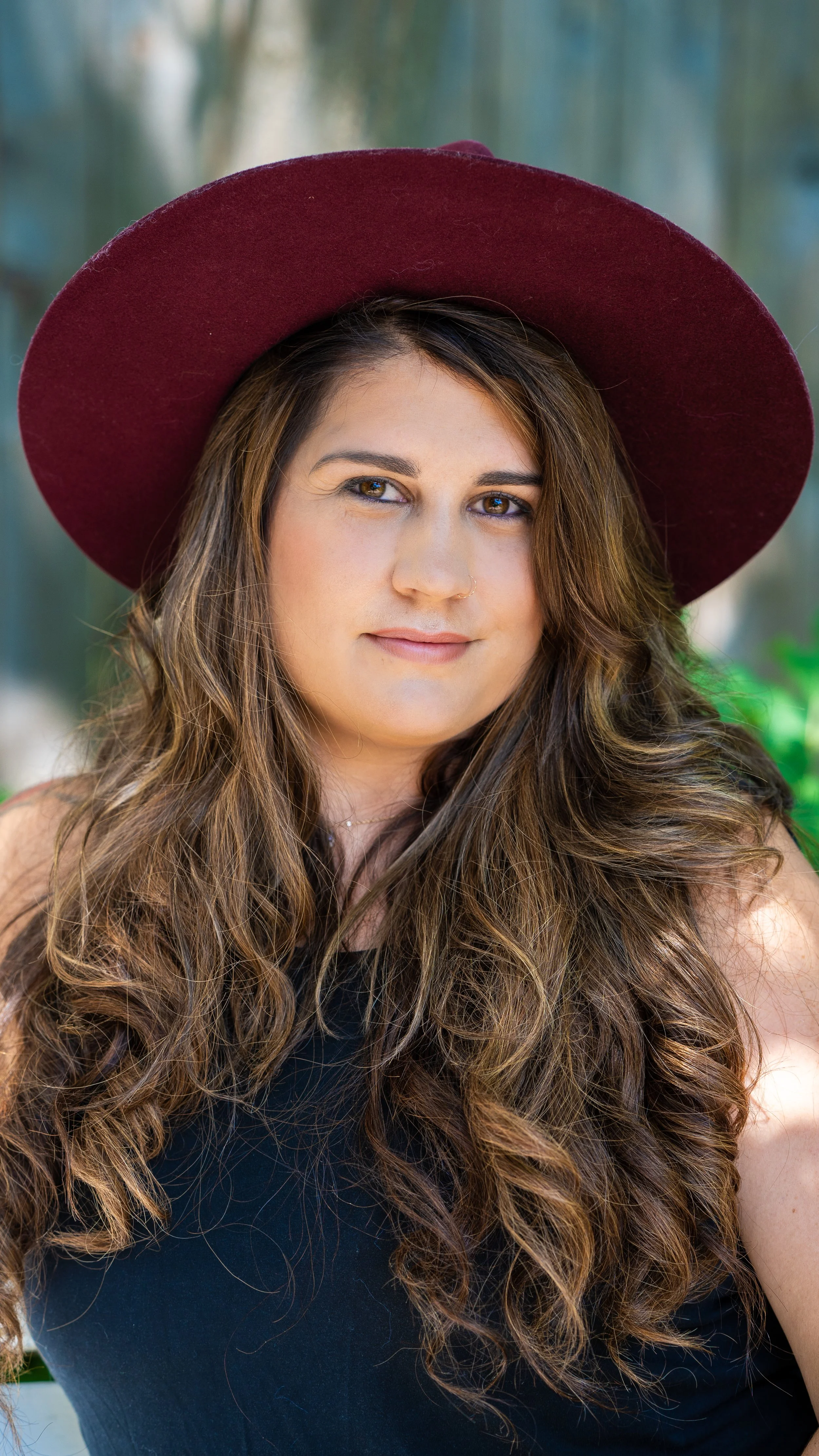 Woman with long hair wearing a large maroon hat and a black top, outdoors with blurred green background.